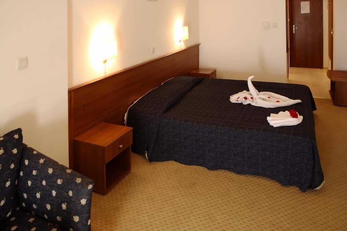 A hotel room with a large bed, a wooden nightstand, a chair, and a wall-mounted lamp.