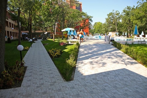 A pathway leading to a pool area with lounge chairs and umbrellas, surrounded by greenery and buildings. - Yunona (Nessebar)