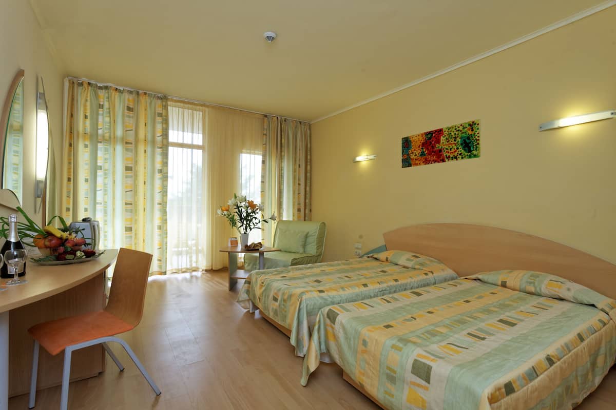 A hotel room with two beds, a desk with a chair, a small table with a vase of flowers, and a colorful painting on the wall.