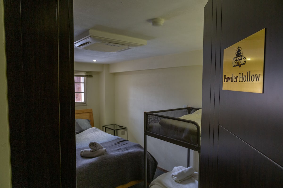 Quarto de hotel com beliche, mesa de cabeceira e janela. Um aviso na parede diz 'Powder Hollow'.