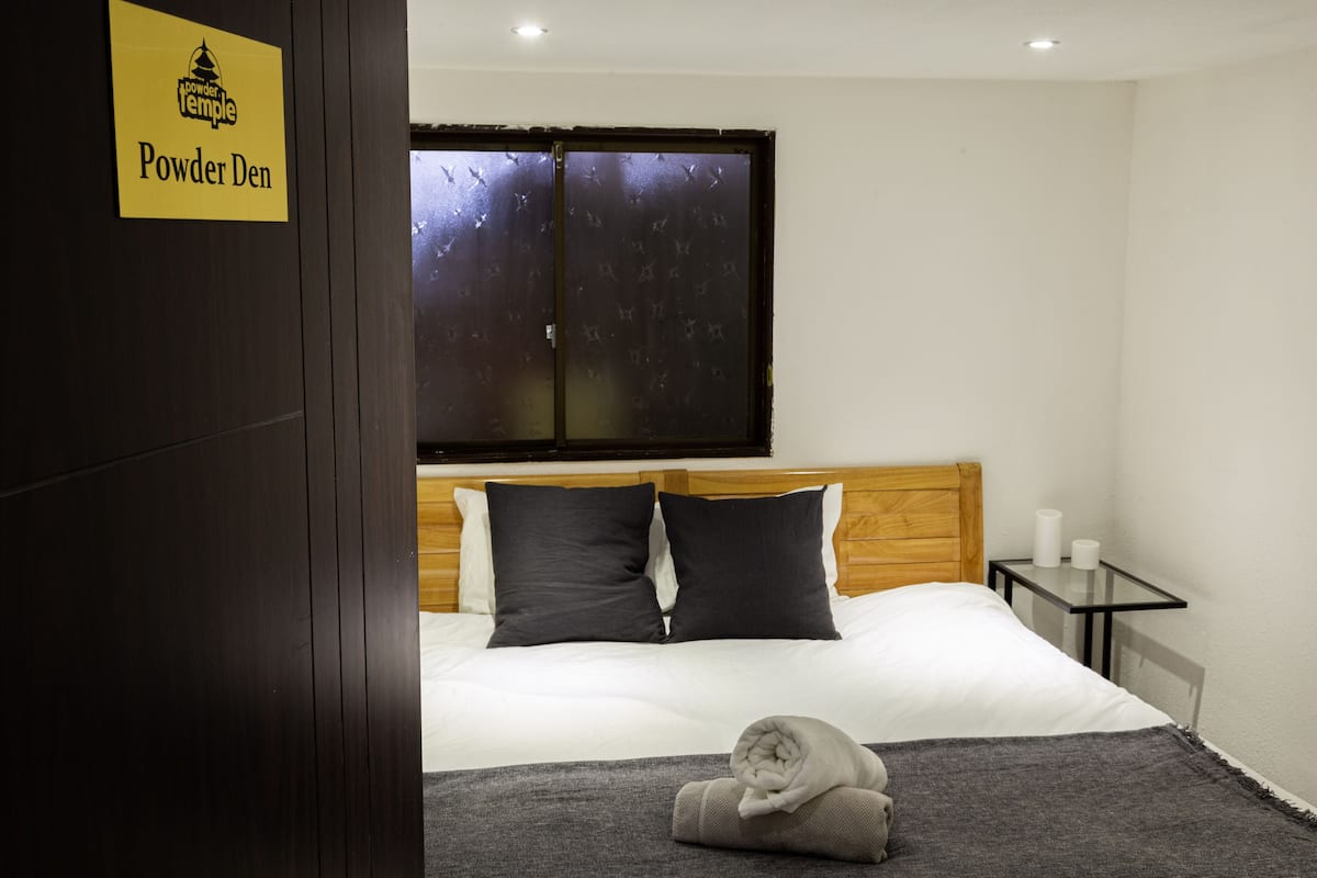 Quarto de hotel com uma cama, mesa de cabeceira, janela com vista e placa indicando o nome do quarto.
