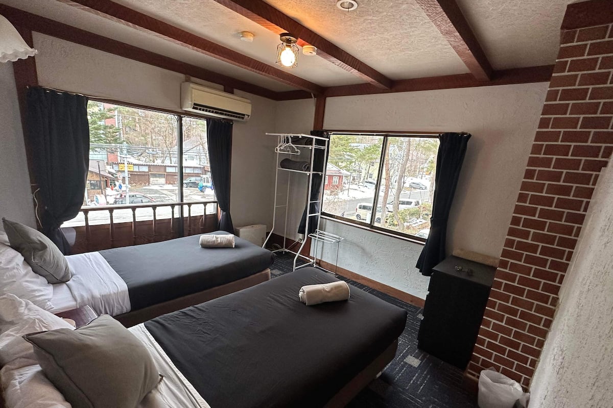Quarto de hotel com duas camas, janela com vista para a rua, cabideiro e parede de tijolos.