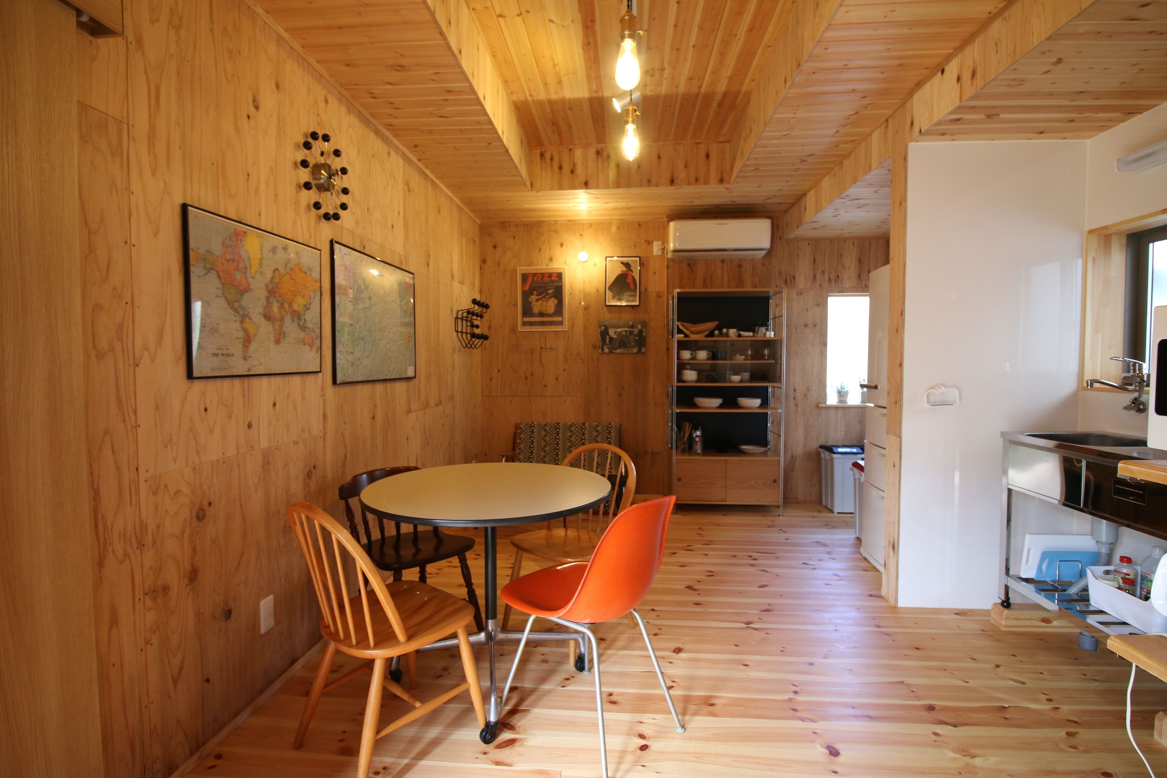 A wooden interior with a round dining table, two wooden chairs, and a kitchen area with shelves and a sink.