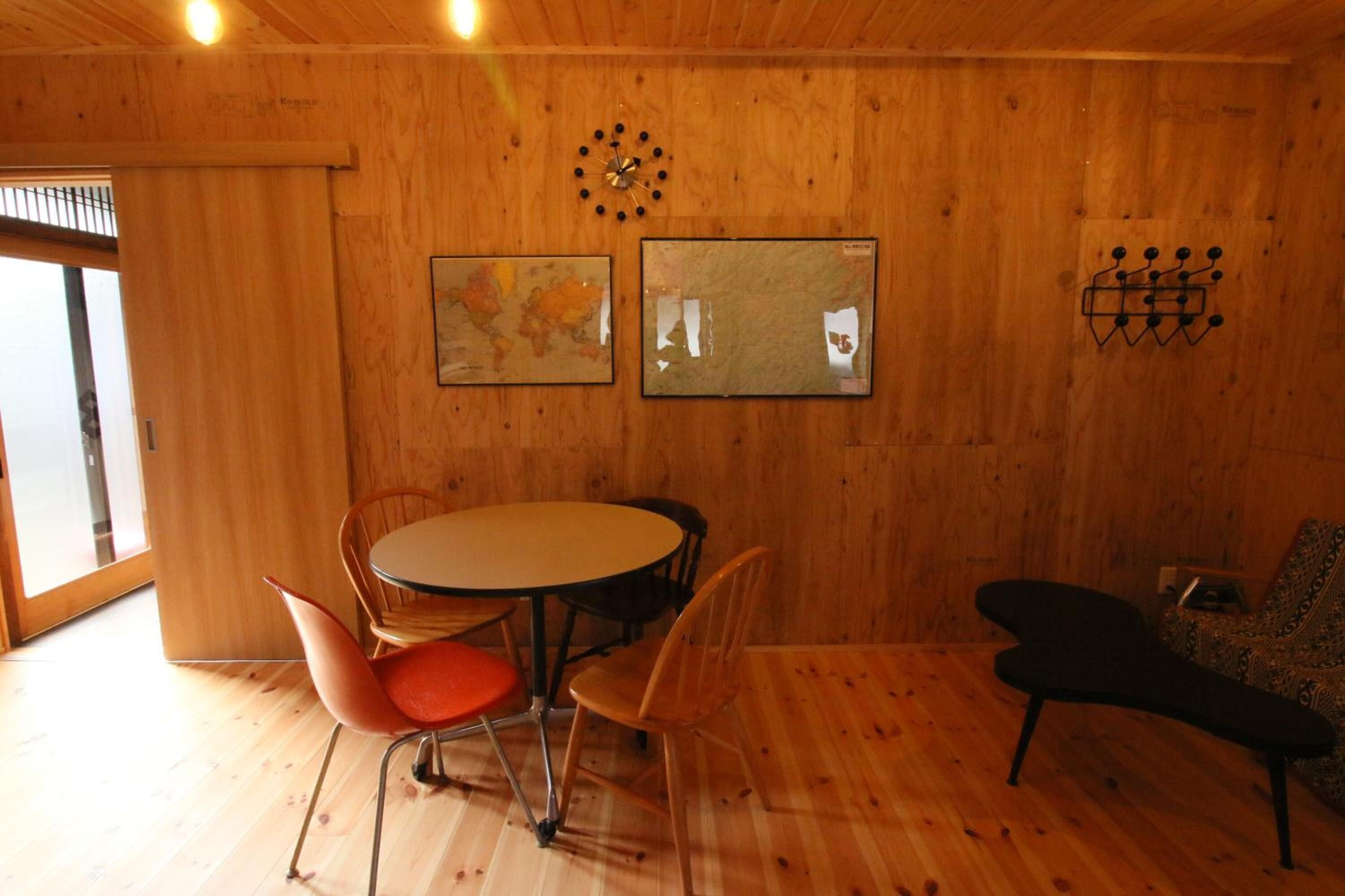 A wooden interior with a round table and chairs, a clock, and two framed pictures on the wall.