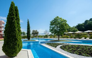 Una piscina con varias pistas, rodeada de setos y árboles cuidadosamente podados, y un edificio con balcones al fondo.