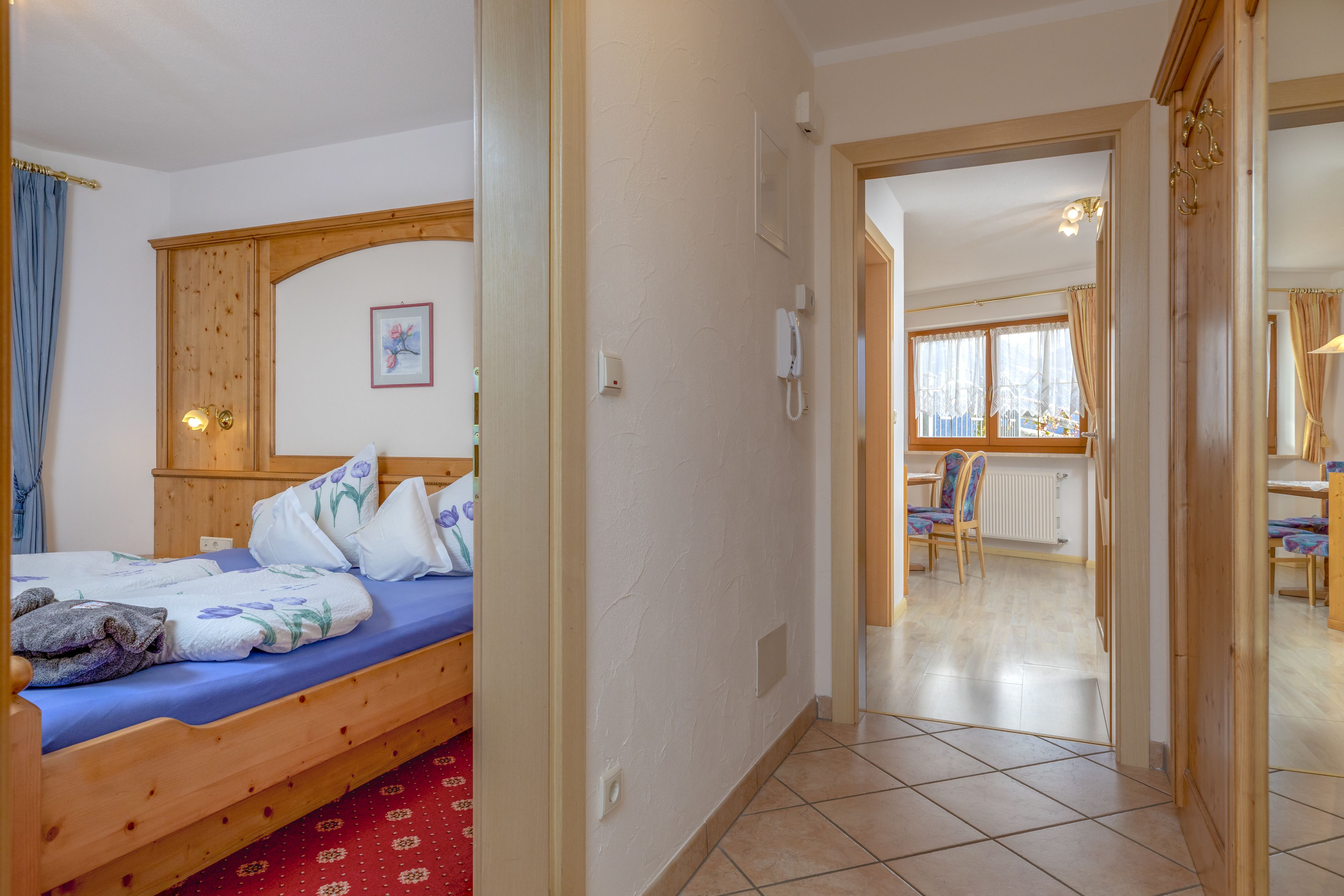 Quarto com cama de madeira, carpete vermelho estampado e vista para uma sala de jantar com cadeiras e mesa.