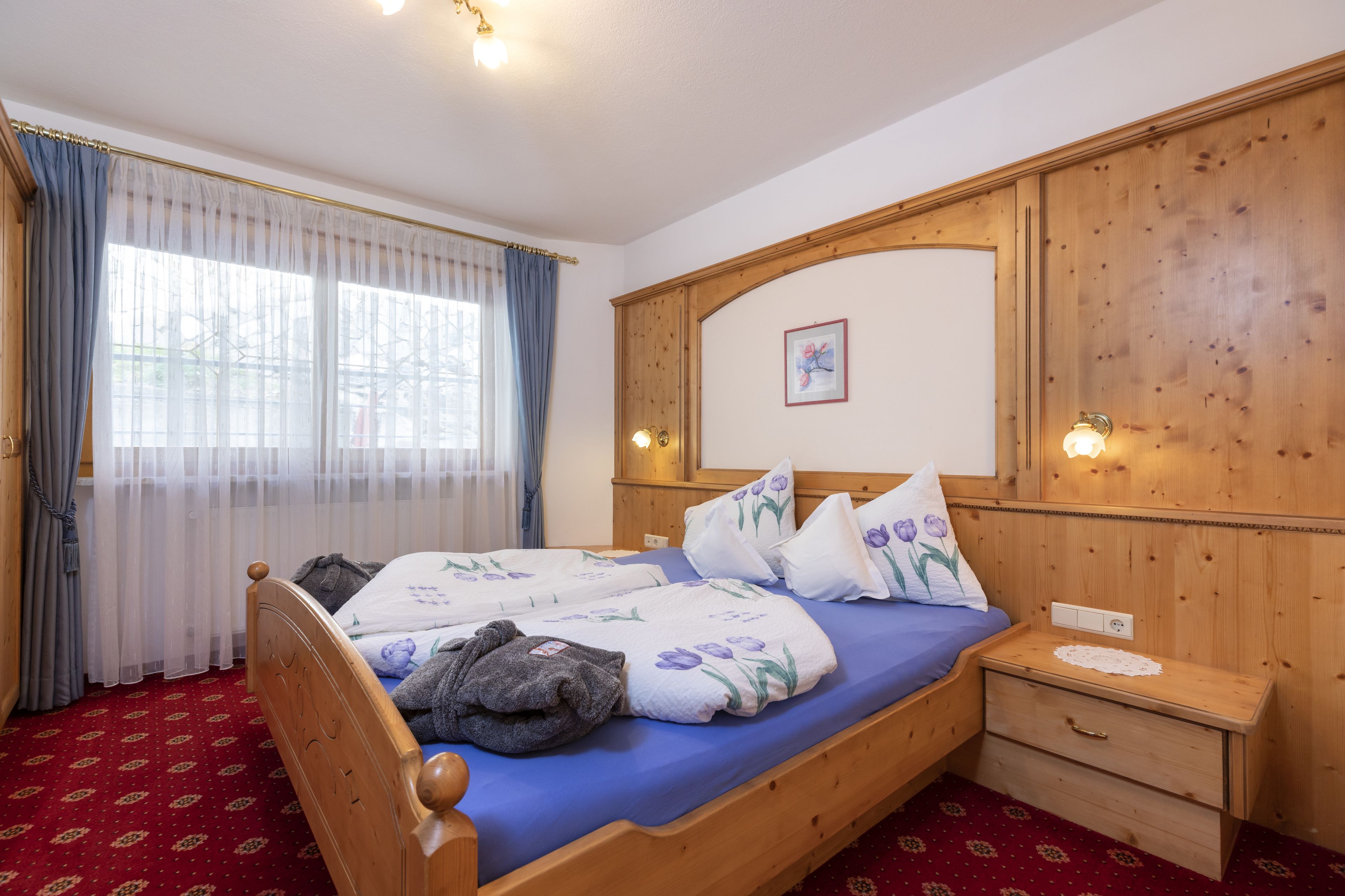 Een houten bed met een blauwe matras, bloemenkussens en een nachtkastje met een lade.