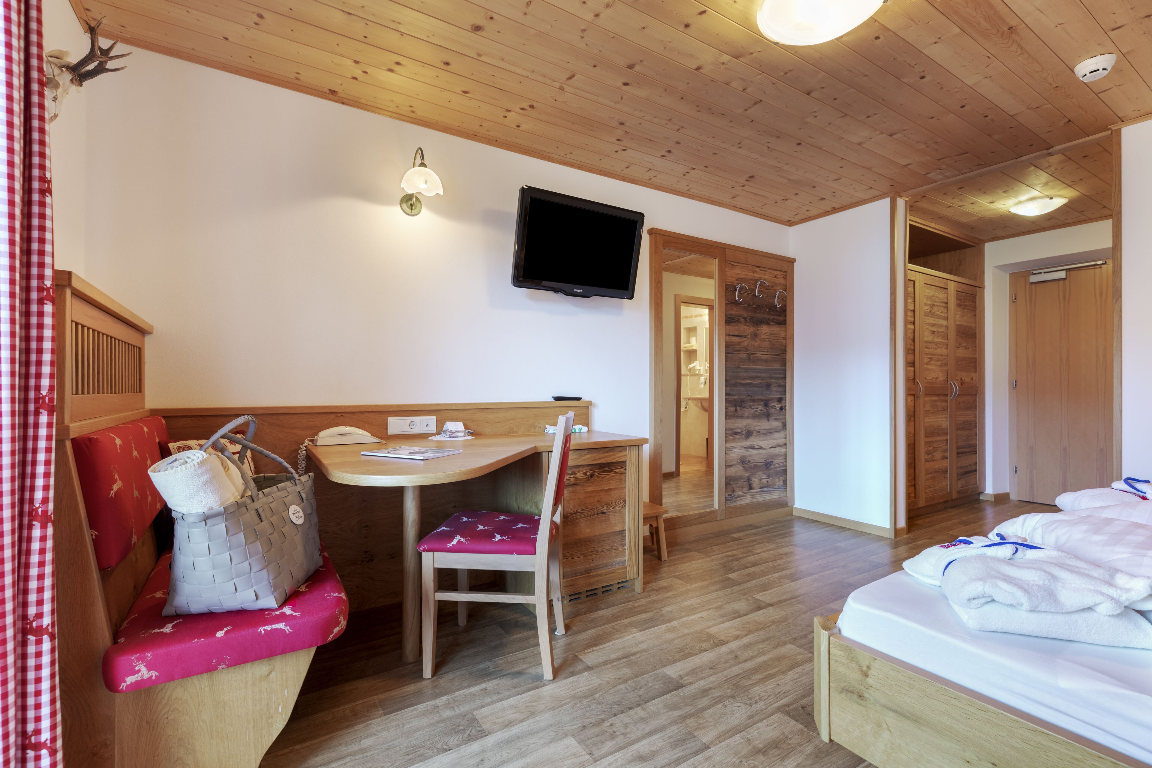 A cozy room with a wooden ceiling, a bed with white linens, a desk with a chair, a television mounted on the wall, and a wooden wardrobe.