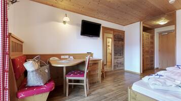 A cozy room with a wooden ceiling, a bed with white linens, a desk with a chair, a television mounted on the wall, and a wooden wardrobe.
