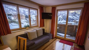Una sala de estar acogedora con un sofá, un televisor montado en la pared y grandes ventanas que ofrecen vistas a un paisaje montañoso nevado.