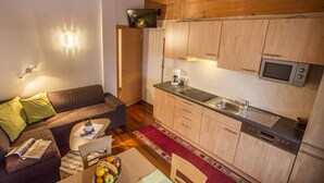 Un espacio habitable compacto con kitchenette, un sofá con cojines, una mesita con una planta y una revista, y un frutero.