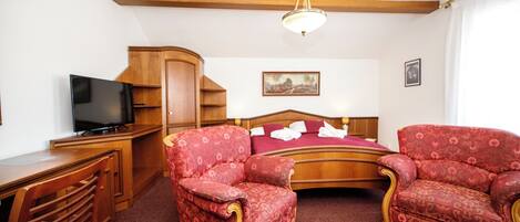 A hotel room with a wooden desk, a television, two red upholstered chairs, a bed with a wooden headboard, and a painting on the wall.