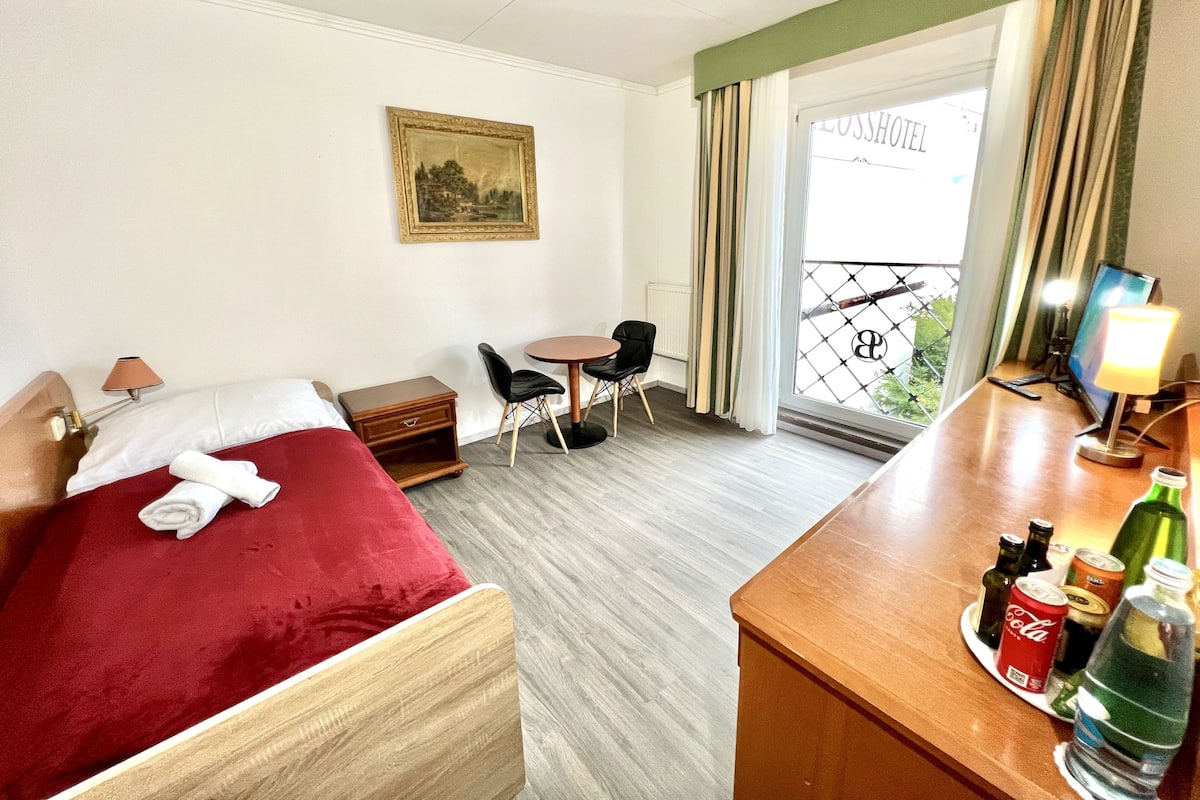 Une chambre d’hôtel avec un lit, un bureau, une chaise, une petite table, un tableau au mur et une fenêtre avec des rideaux.
