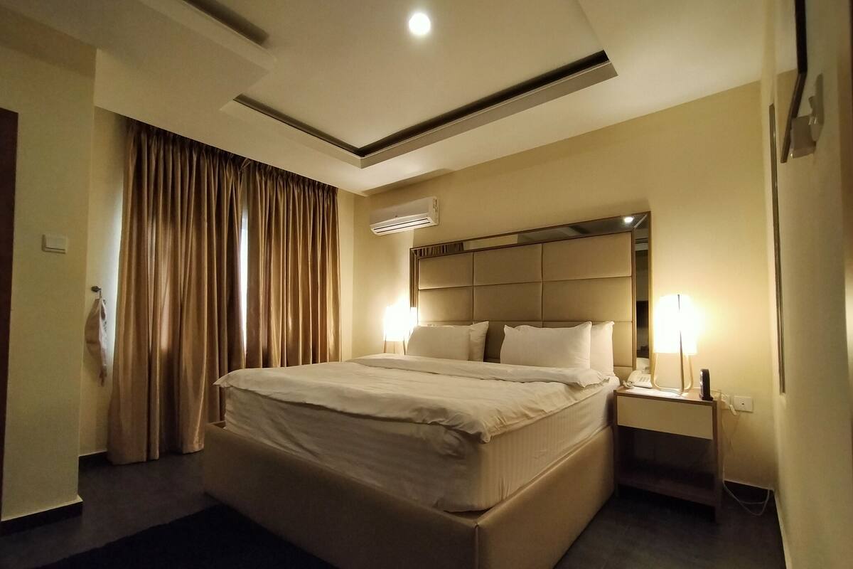 A hotel room with a large bed, a nightstand with a lamp, a ceiling light, and a window with curtains.