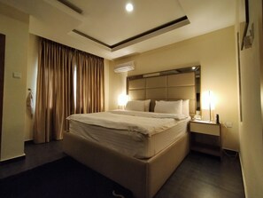 A hotel room with a large bed, a nightstand with a lamp, a ceiling light, and a window with curtains. - Iris Hotel (Abuja)