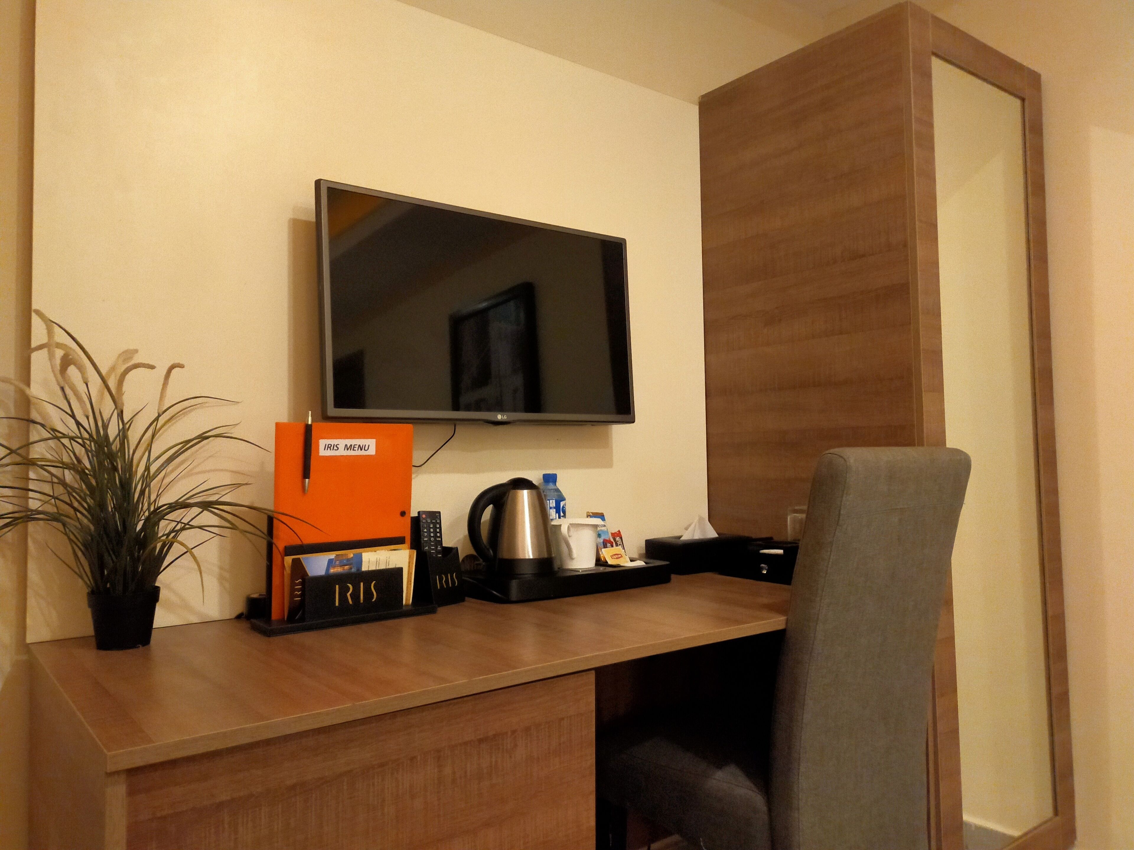 A hotel room with a wooden desk, a television mounted on the wall, a plant, and a seating area with a chair.
