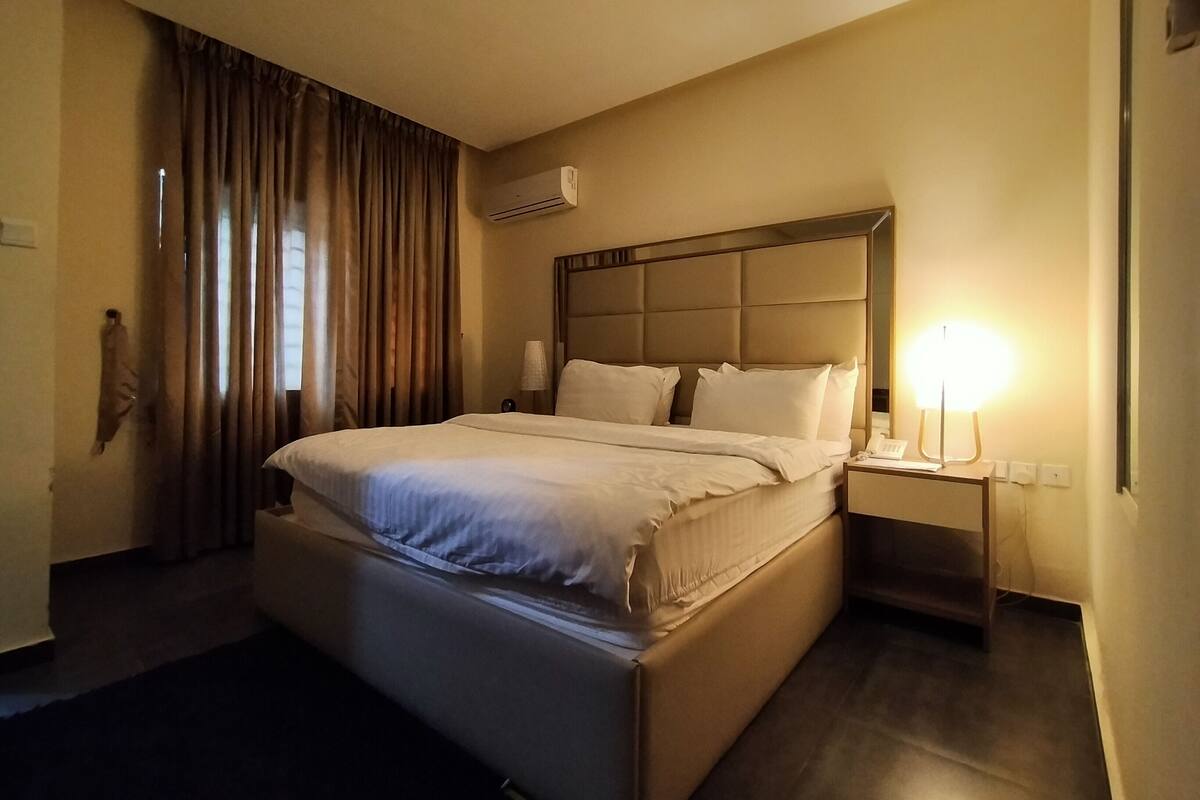 A hotel room with a large bed, a nightstand, a lamp, and an air conditioning unit on the wall.
