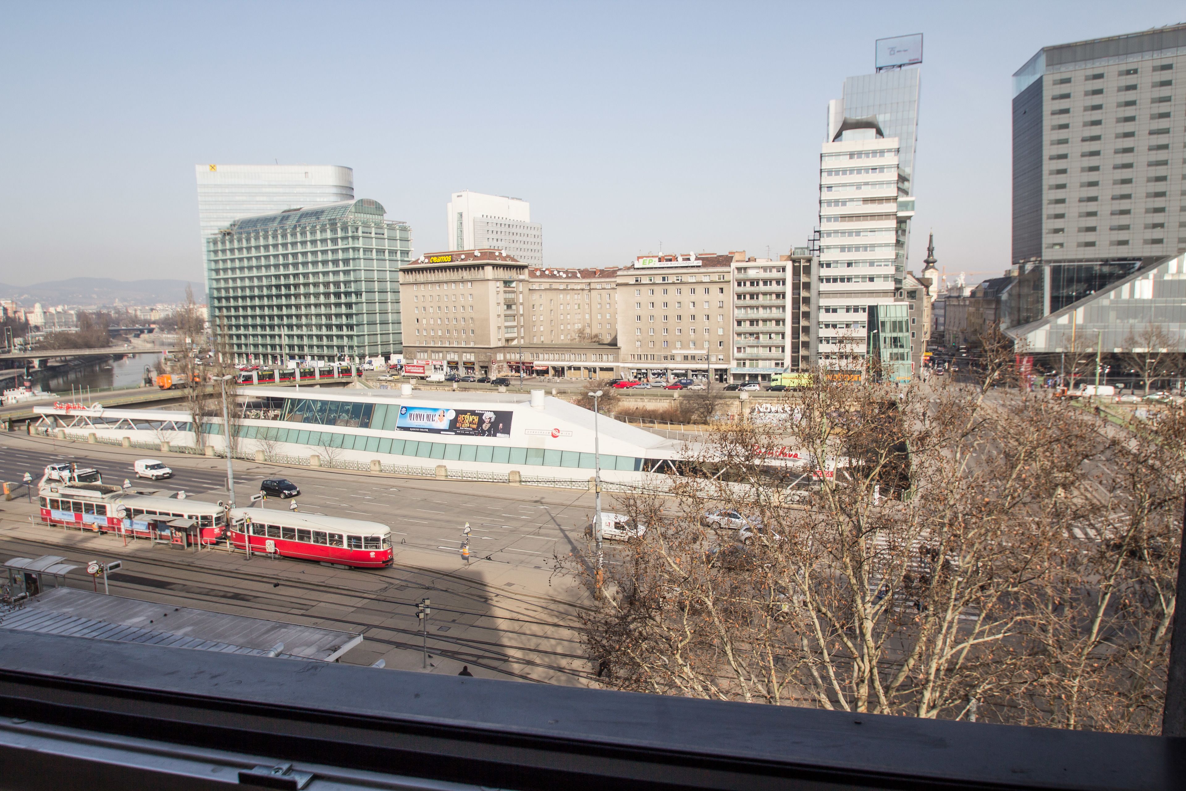 Stadtbild mit einem markanten Gebäude, einer großen Werbetafel, einer Straßenbahntrasse und einer Mischung aus moderner und traditioneller Architektur.