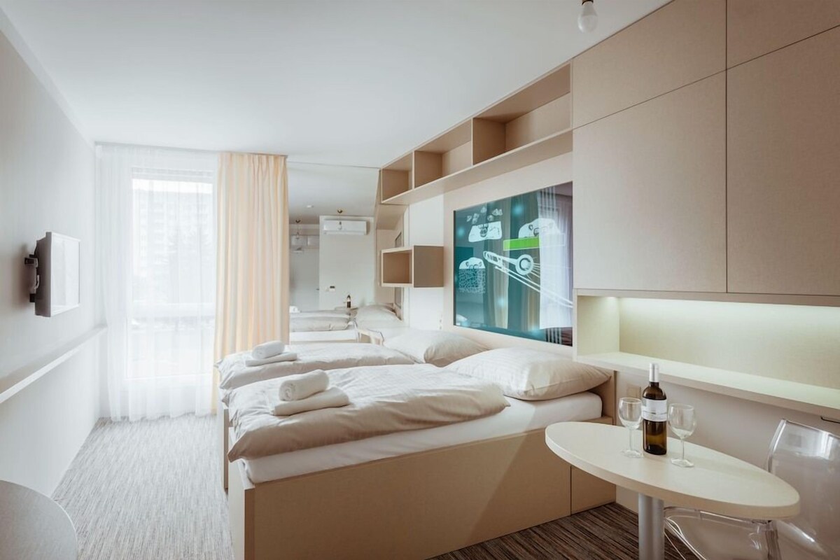 A hotel room with a bed, a small table with glasses and a bottle, and a large window with curtains.