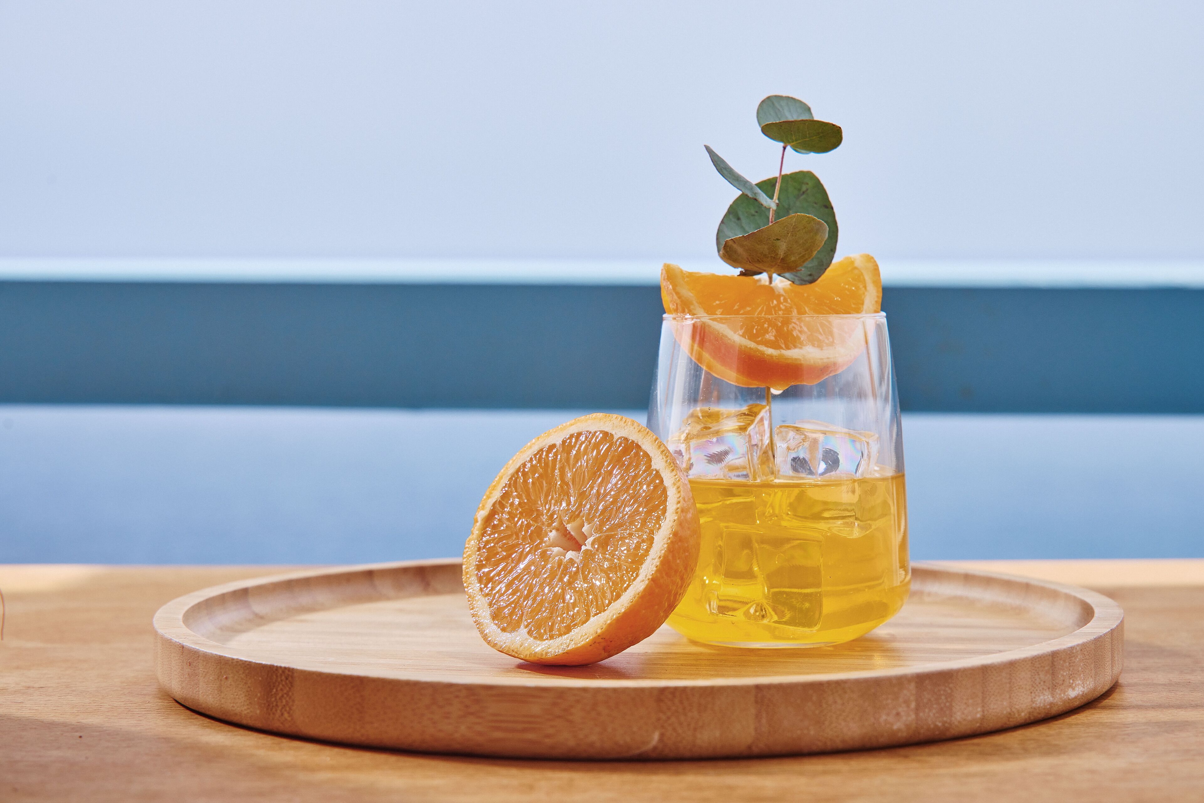 A glass of yellow liquid with a slice of orange and a leaf garnish on a wooden tray.