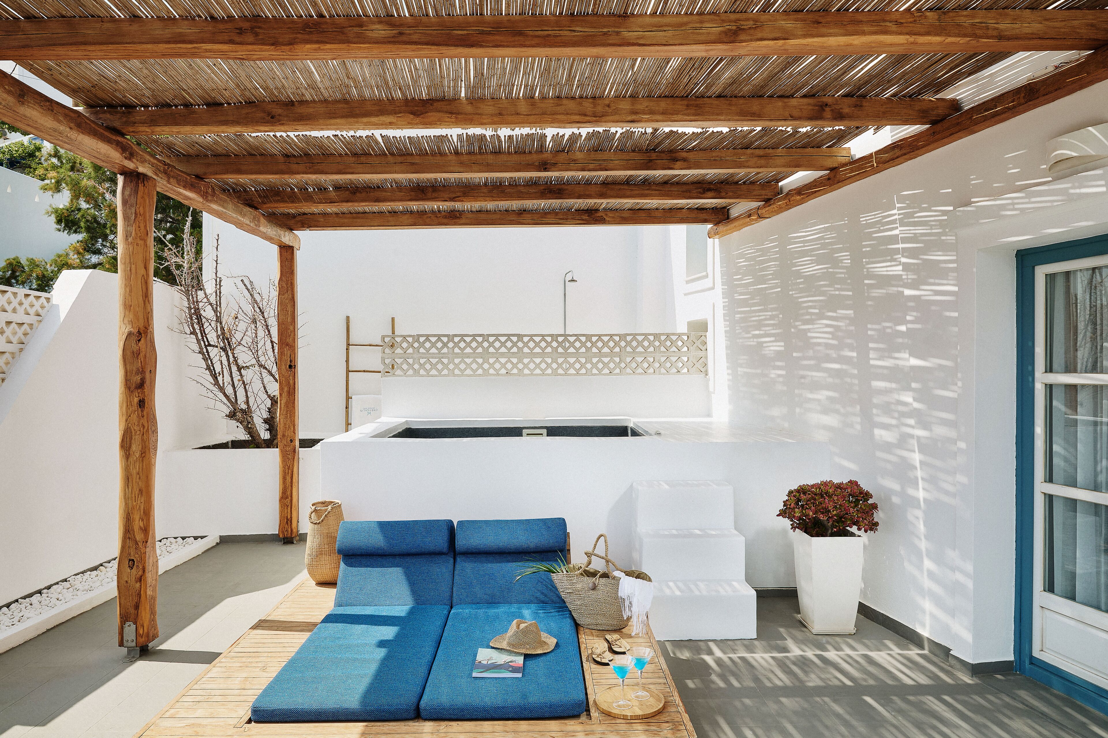 A covered outdoor seating area with a wooden pergola, a blue sofa, a white chair, and a small table with a plant and a hat.