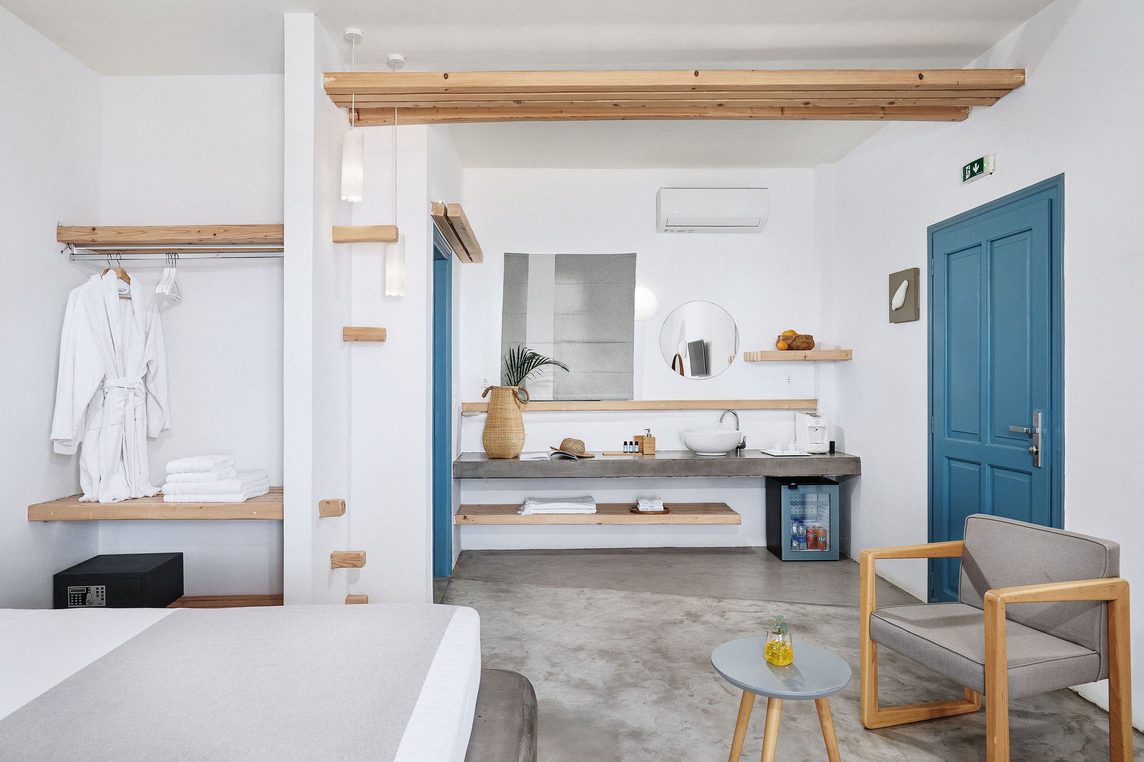 A modern hotel room with a bed, a seating area, a small table, a bathroom with a sink, and a blue door.
