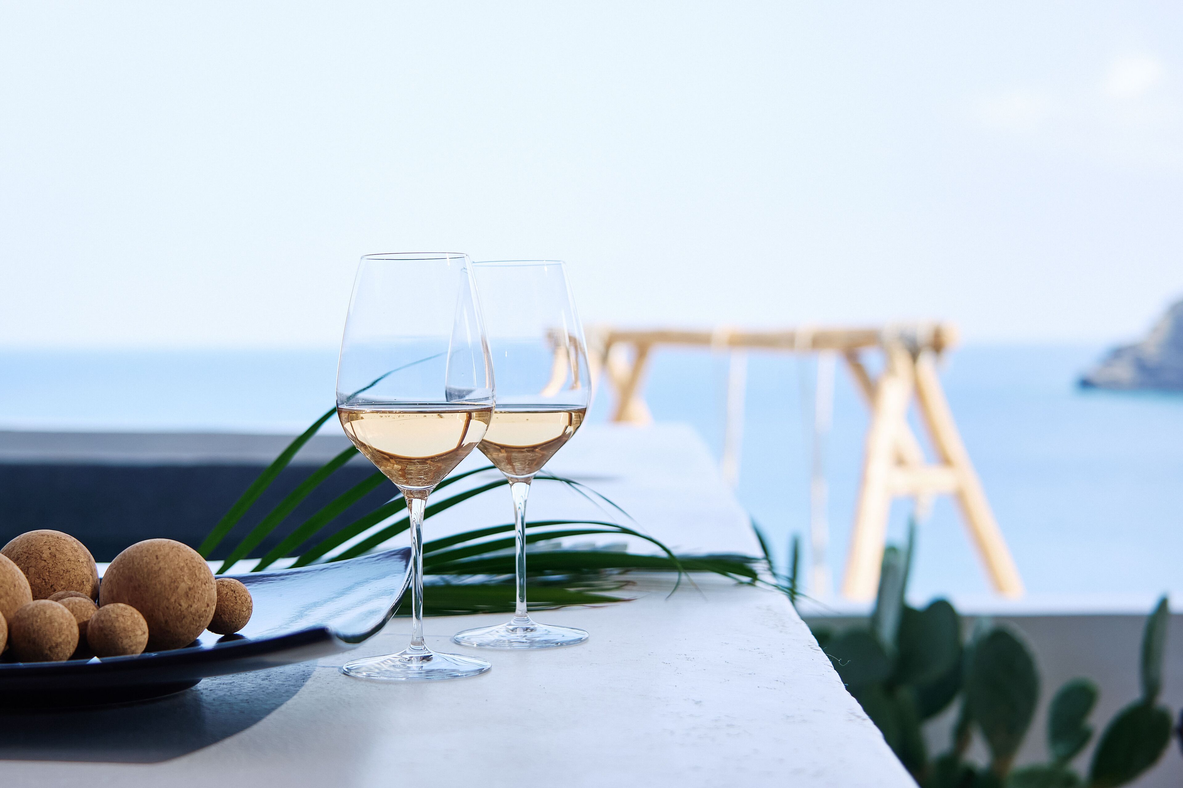 Dos copas de vino blanco sobre una mesa, con un plato de objetos redondos y marrones, posiblemente nueces, y una silla de madera al fondo con vistas al mar.