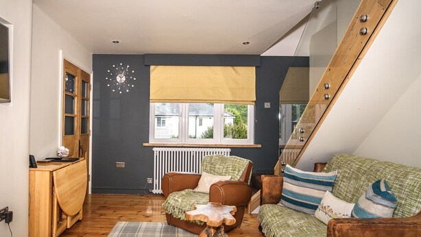 Una habitación acogedora con un sofá de cuero marrón, una mesa de centro de madera y una alfombra de cuadros. Se ve una ventana con vistas a casas.