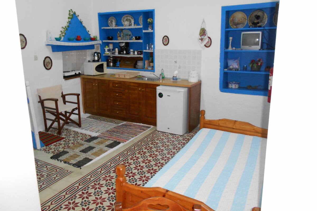 A compact kitchen with wooden cabinets, a small refrigerator, and a microwave. There is a dining area with a chair and a bed with a striped bedspread.