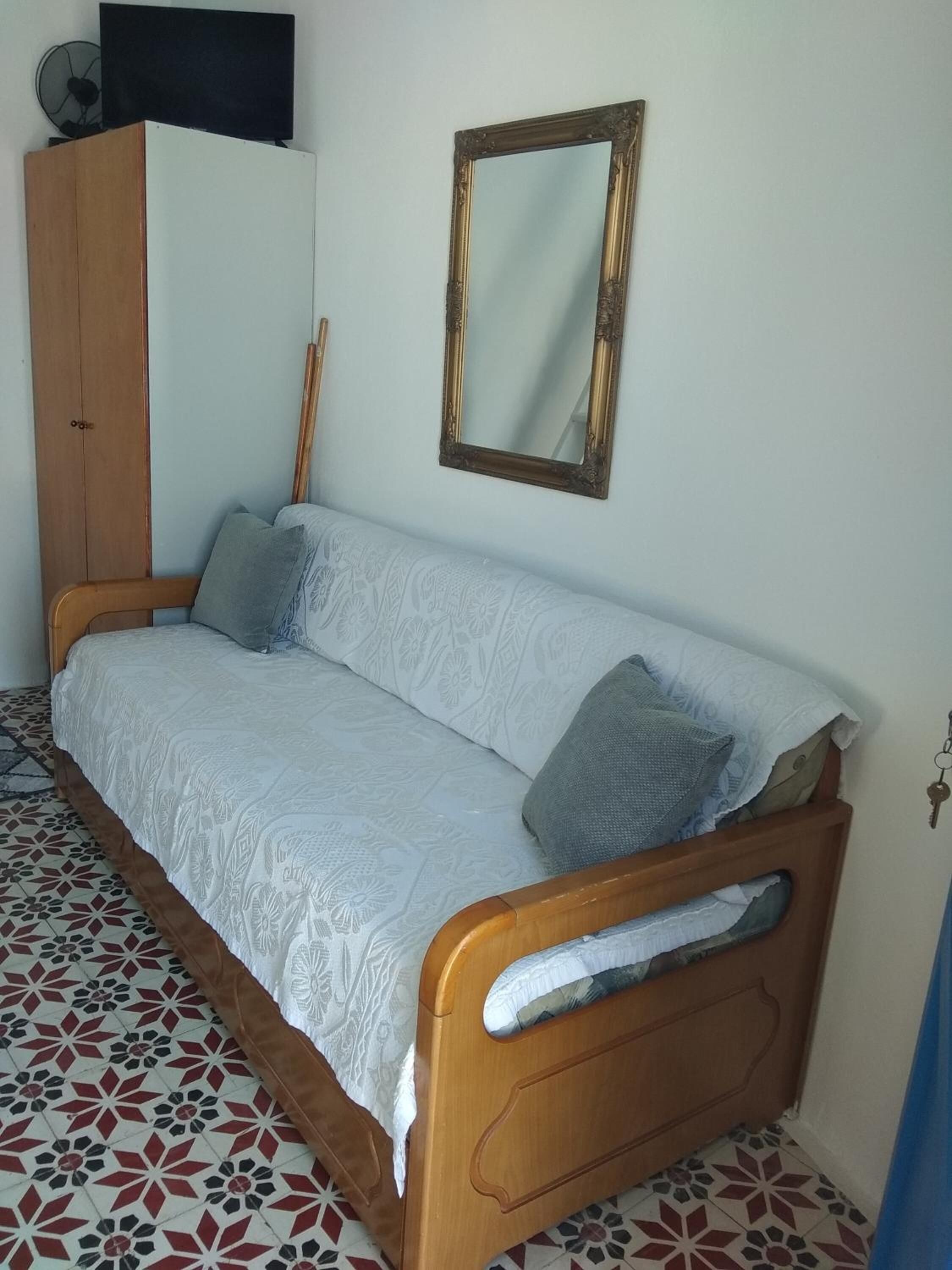 A single-bed room with a wooden frame, a white patterned bedspread, a wooden wardrobe, a mirror, and a television on top of the wardrobe.