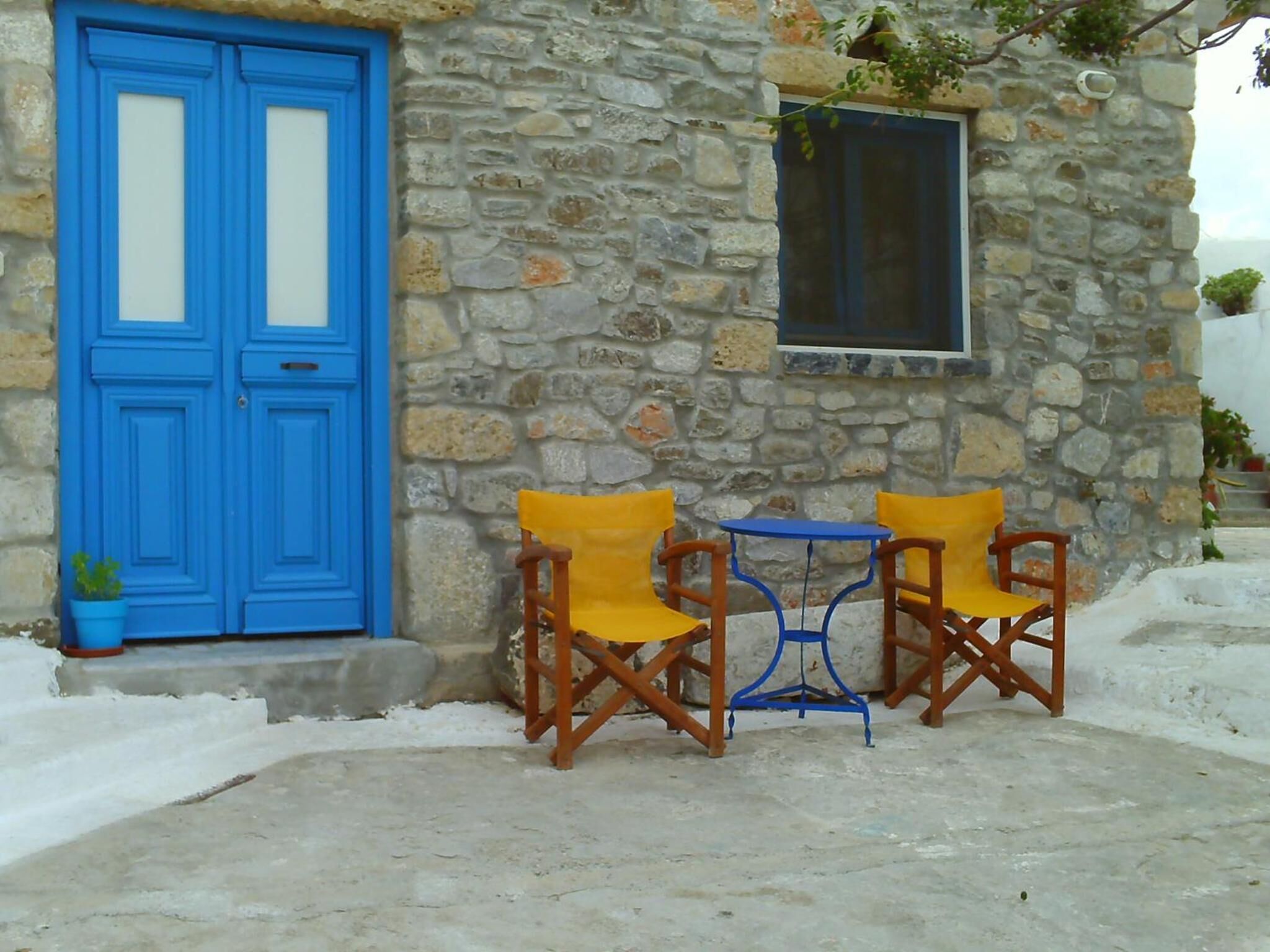 A stone building with a blue door and a small window. There are two wooden chairs with yellow cushions and a blue table in front of the building.