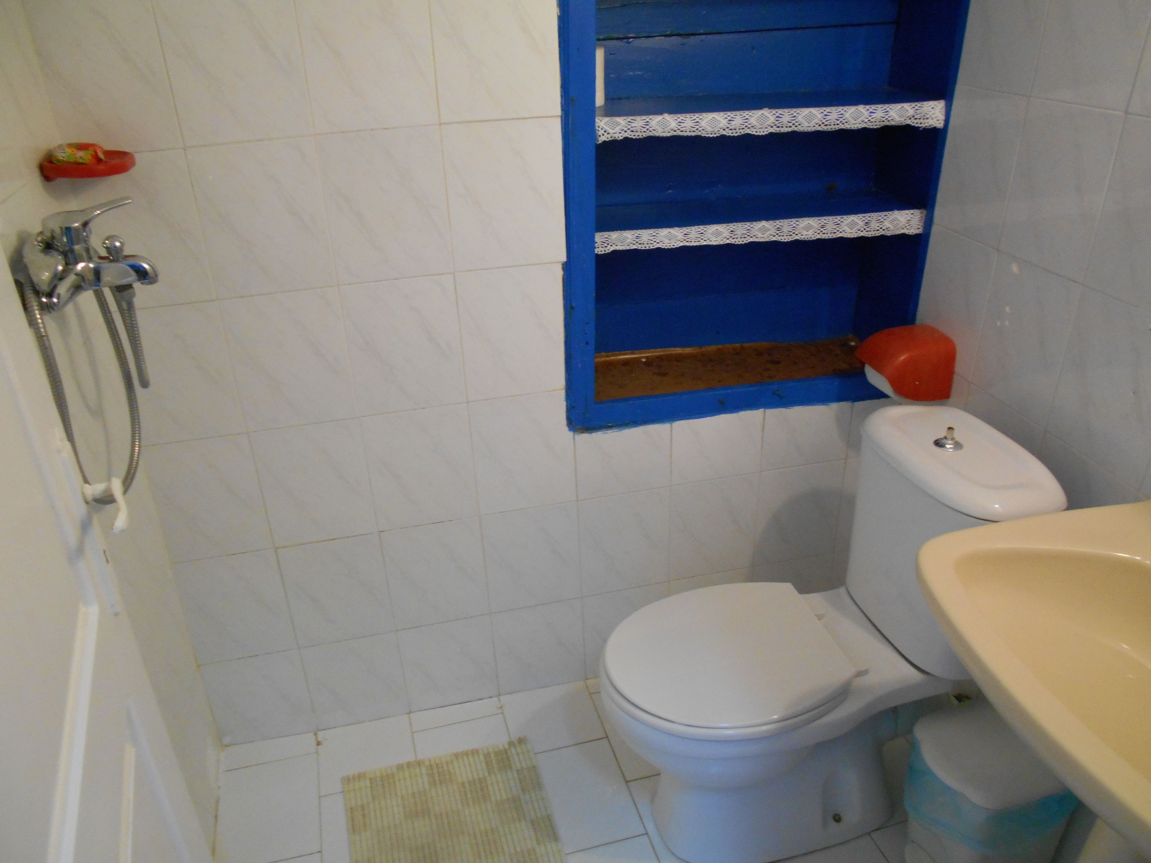 A bathroom with a white toilet, a blue shelf with lace trim, a red object on the floor, and a beige sink.