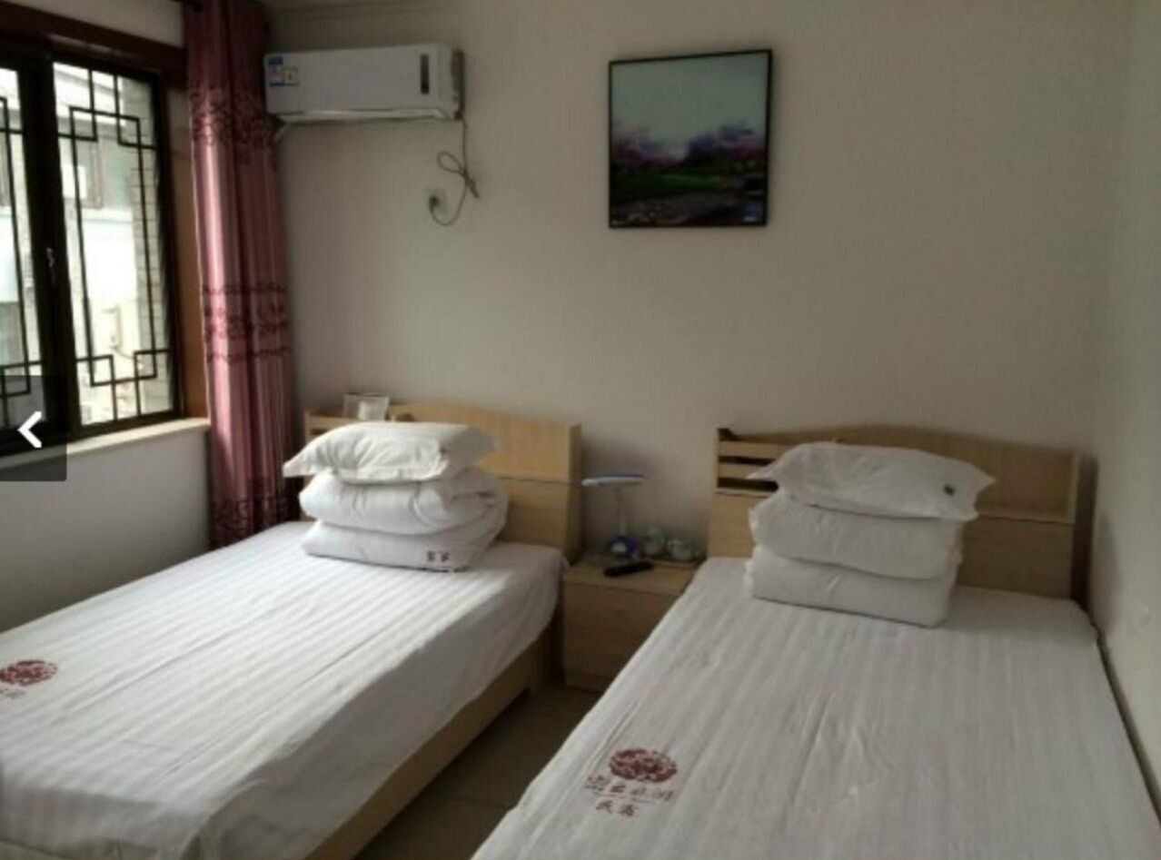 A room with two single beds, each with white bedding and pillows, a small bedside table with a lamp, and a wall-mounted air conditioner.