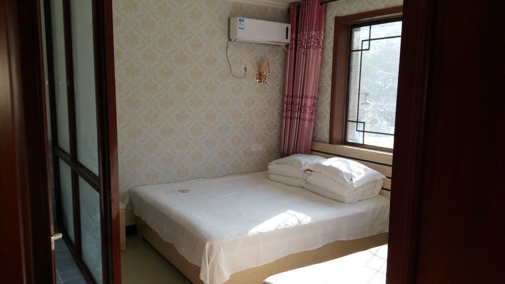 Una cama pequeña y bien arreglada con colchón y almohadas blancos, una ventana con vistas a árboles, y un equipo de aire acondicionado montado en la pared.