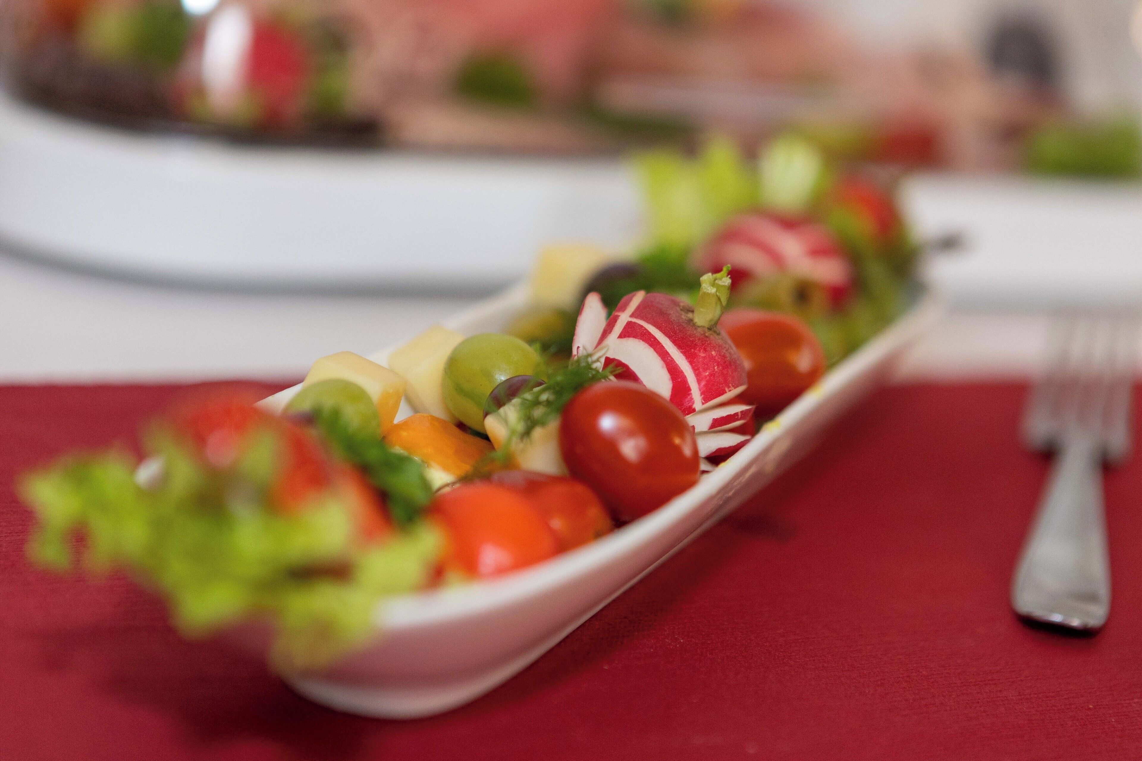 Uma tigela retangular branca cheia de legumes e frutas variadas, incluindo tomates, rabanetes e uvas verdes, sobre uma toalha de mesa vermelha.