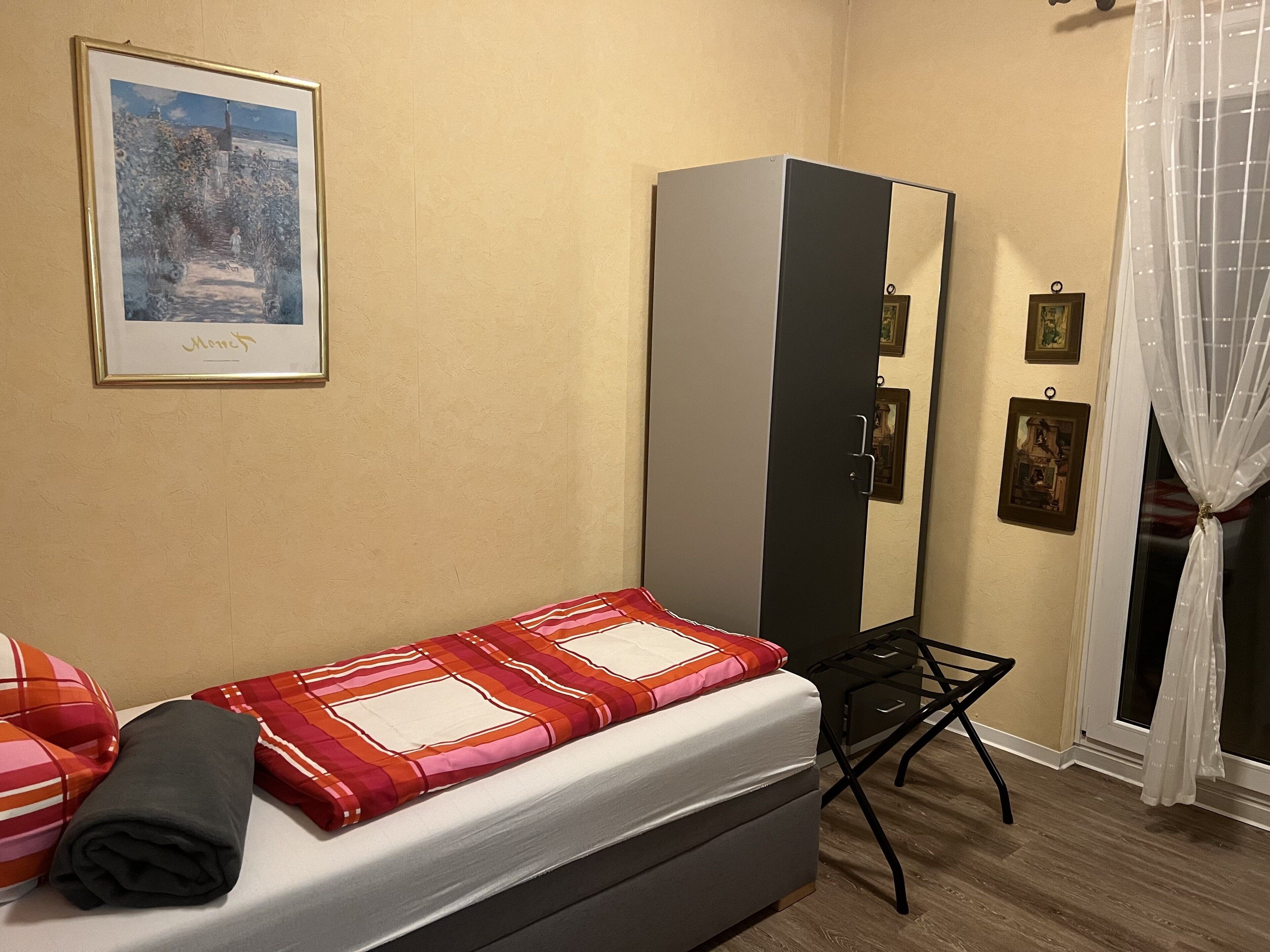 A small bedroom with a bed, a wardrobe, a small table, and a window with curtains.