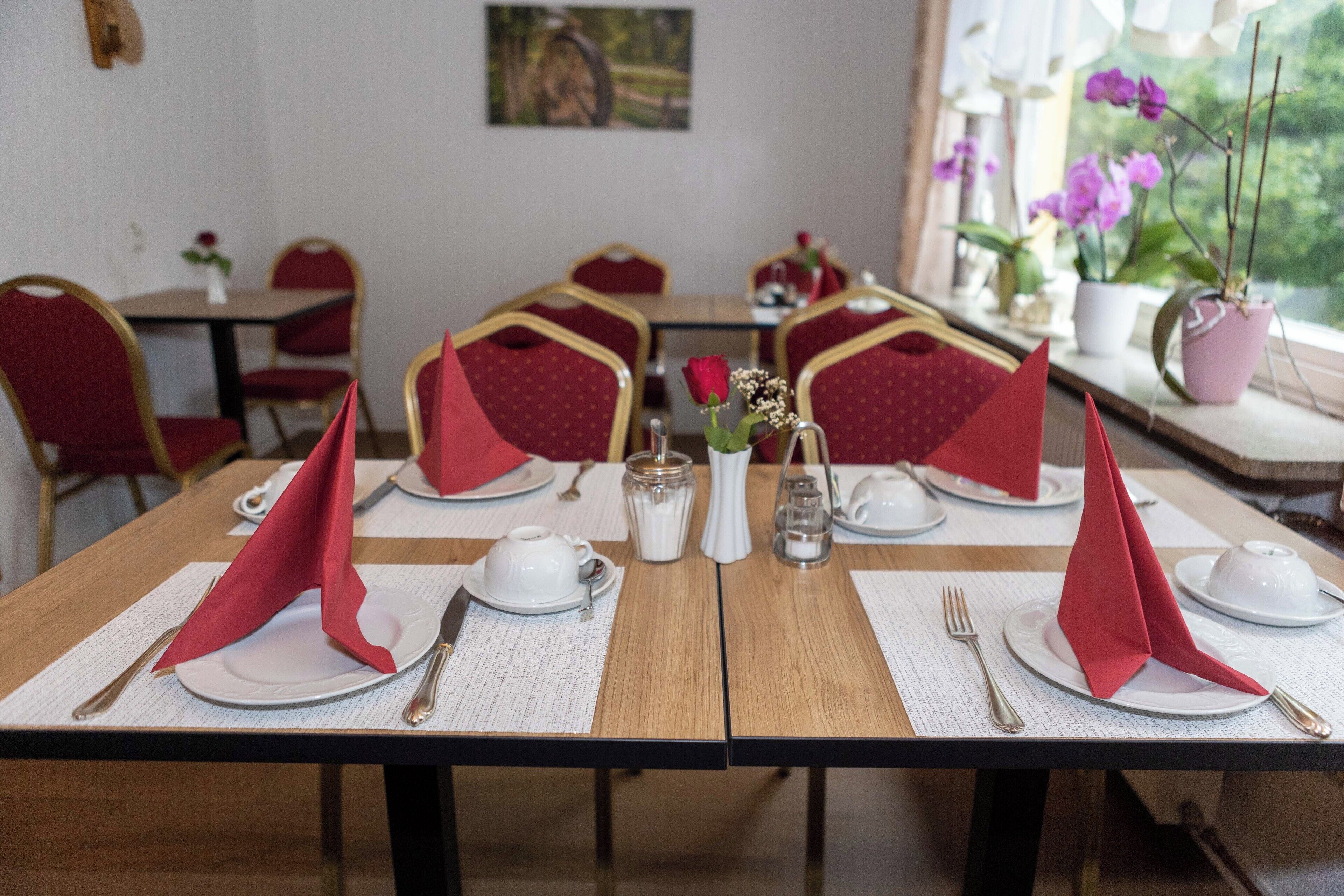 A dining area with tables set for four, each with a red napkin, white plates, and silverware. There are potted plants on the windowsill.