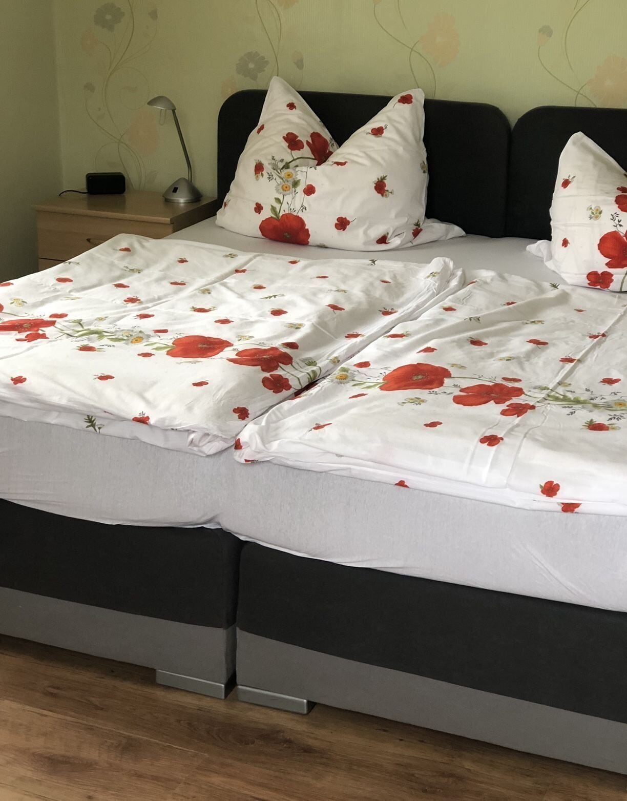 Una cama con colcha estampada de flores, dos almohadas con diseño de flor roja, una mesita de noche con lámpara y un mueble auxiliar con cargador de teléfono.