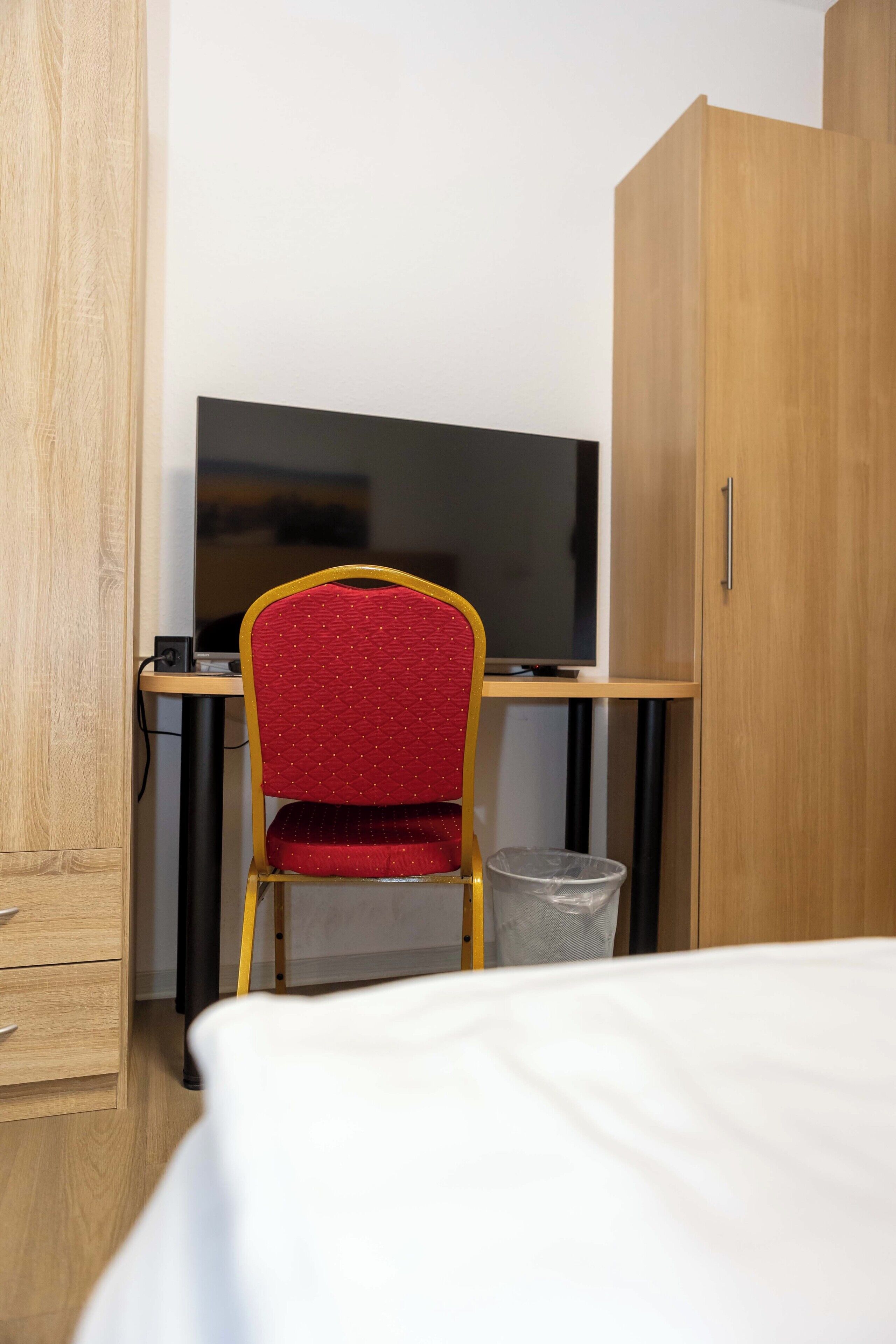Ein Hotelzimmer mit einem Bett, einem Schreibtisch mit Computer, einem roten Sessel, einem Holzschrank und einem Mülleimer.