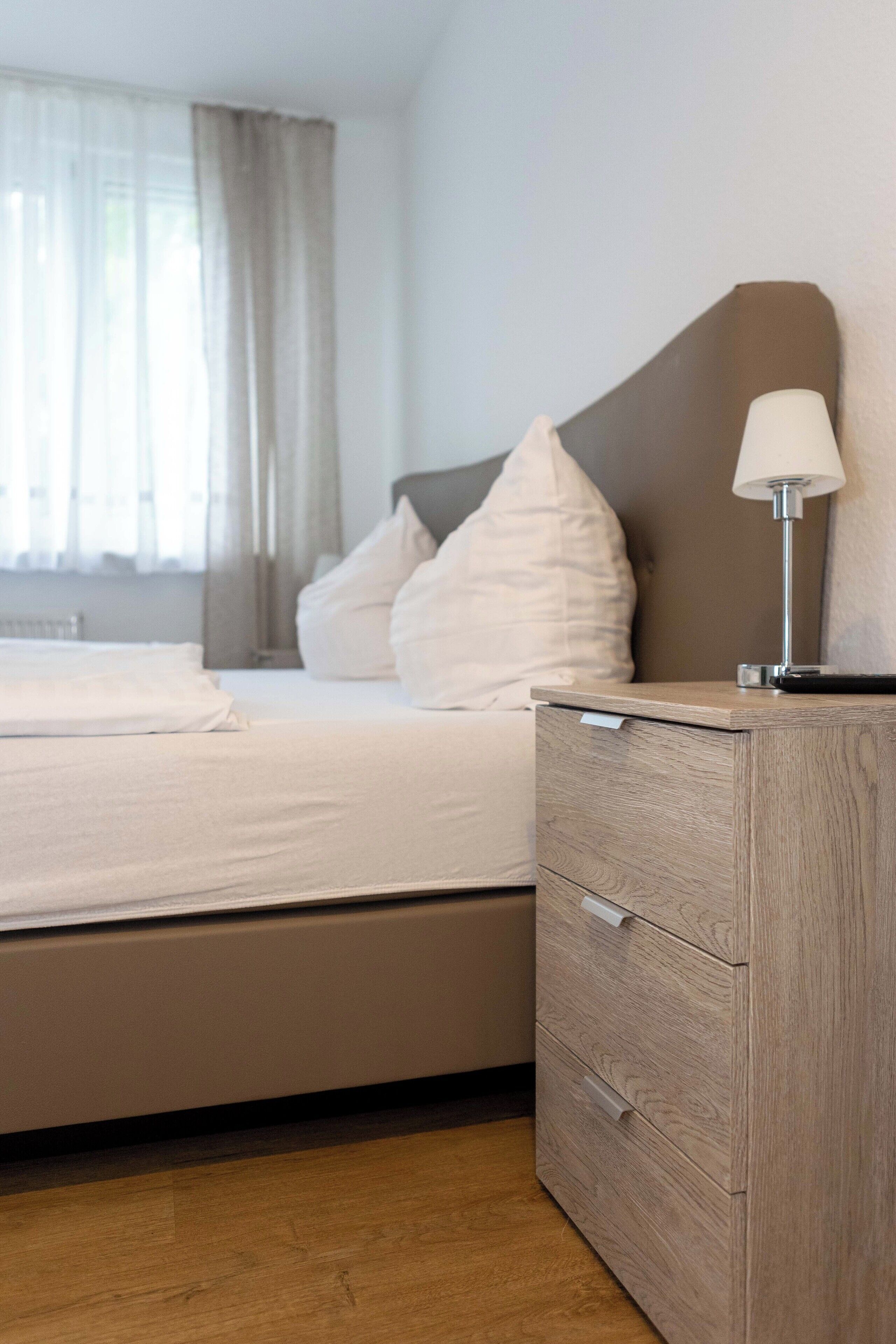 A neatly made bed with white linens, a bedside lamp, and a wooden nightstand with drawers.