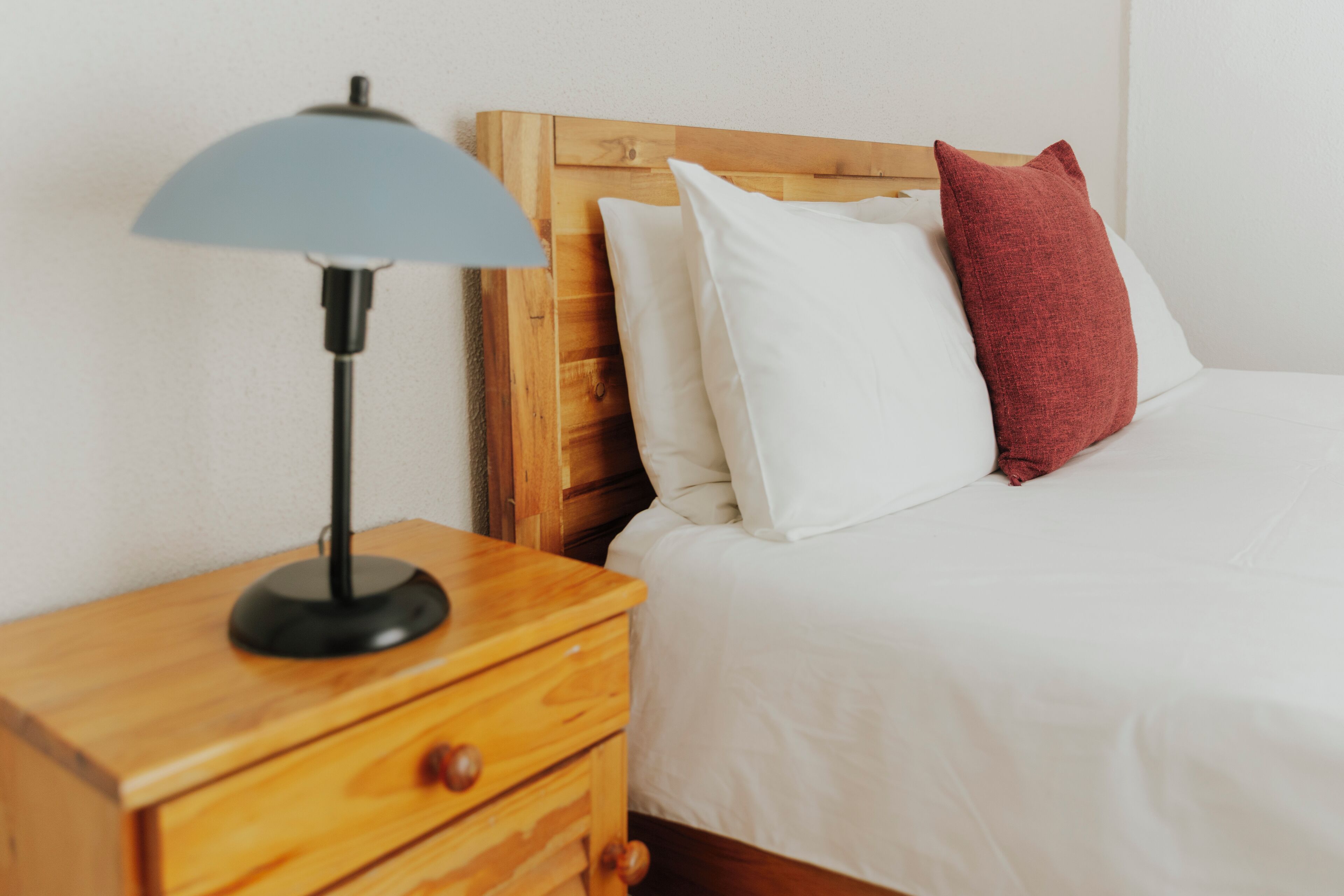 A neatly made bed with white and red pillows, a wooden nightstand with a lamp, and a wooden headboard.