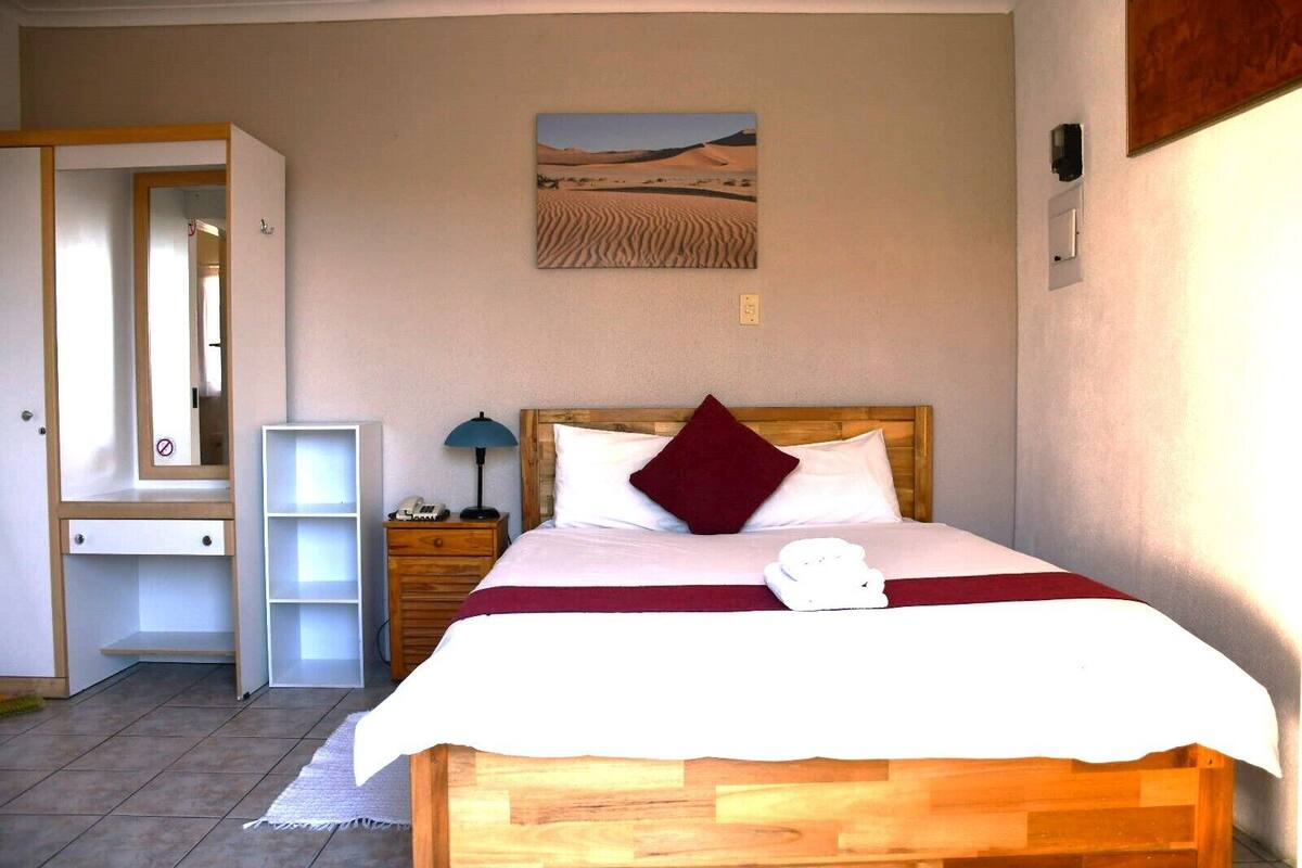 A bedroom with a wooden bed, white bedding, a red pillow, a nightstand with a lamp, a small shelf, and a painting of a desert landscape.