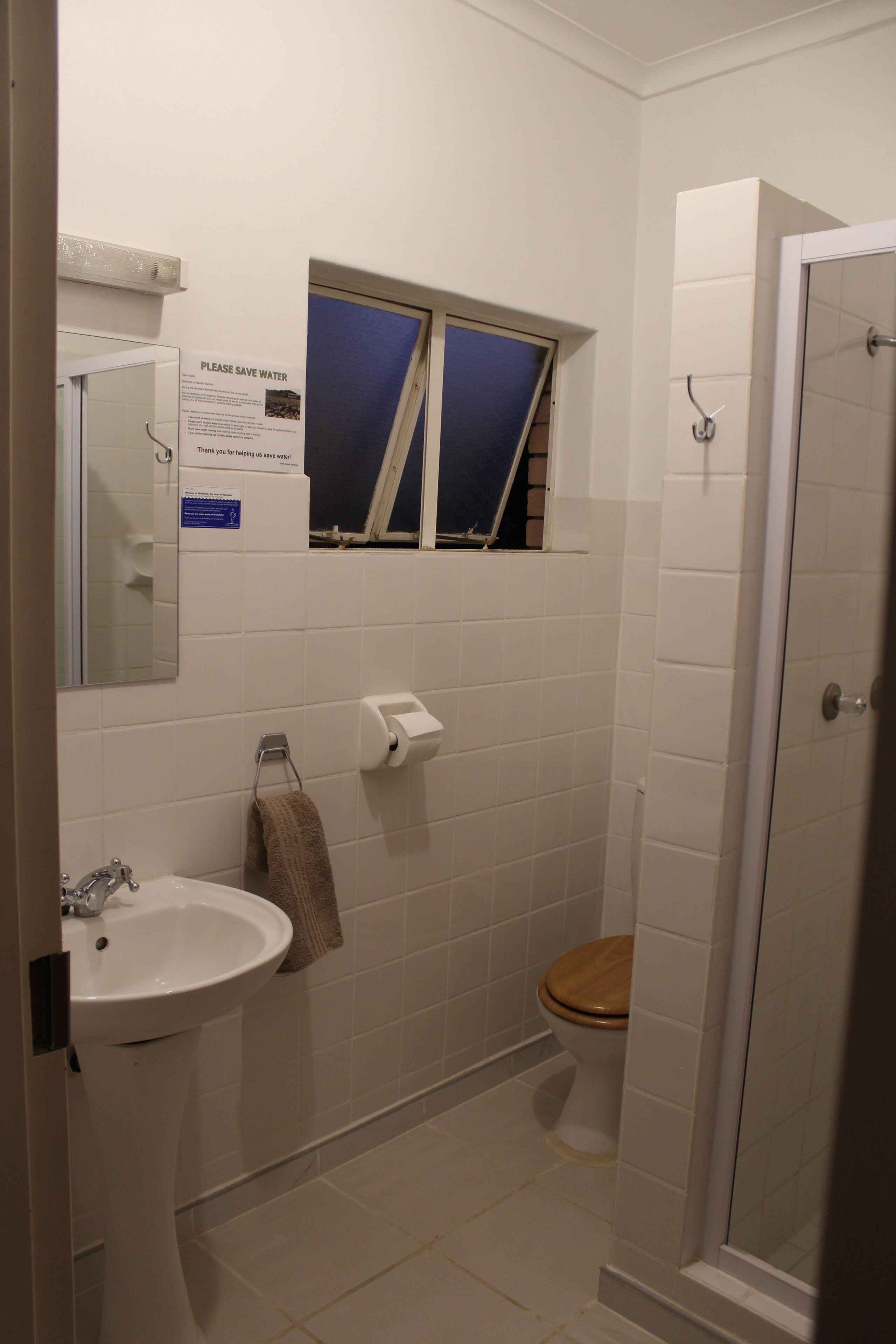 A small bathroom with a sink, toilet, and shower. There is a sign on the wall that says "Please Save Water."