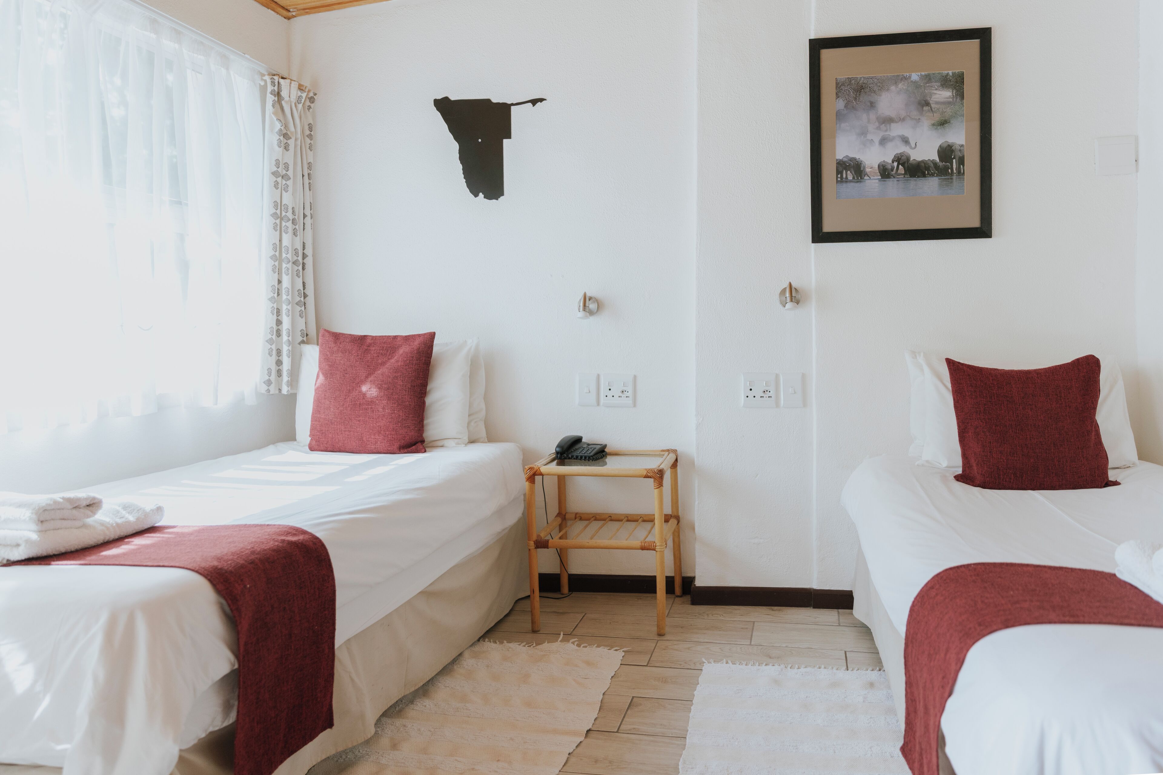 Quarto de hotel com duas camas de solteiro, uma pequena mesa de madeira, uma cabeça de vaca preta fixada na parede e um quadro com elefantes.