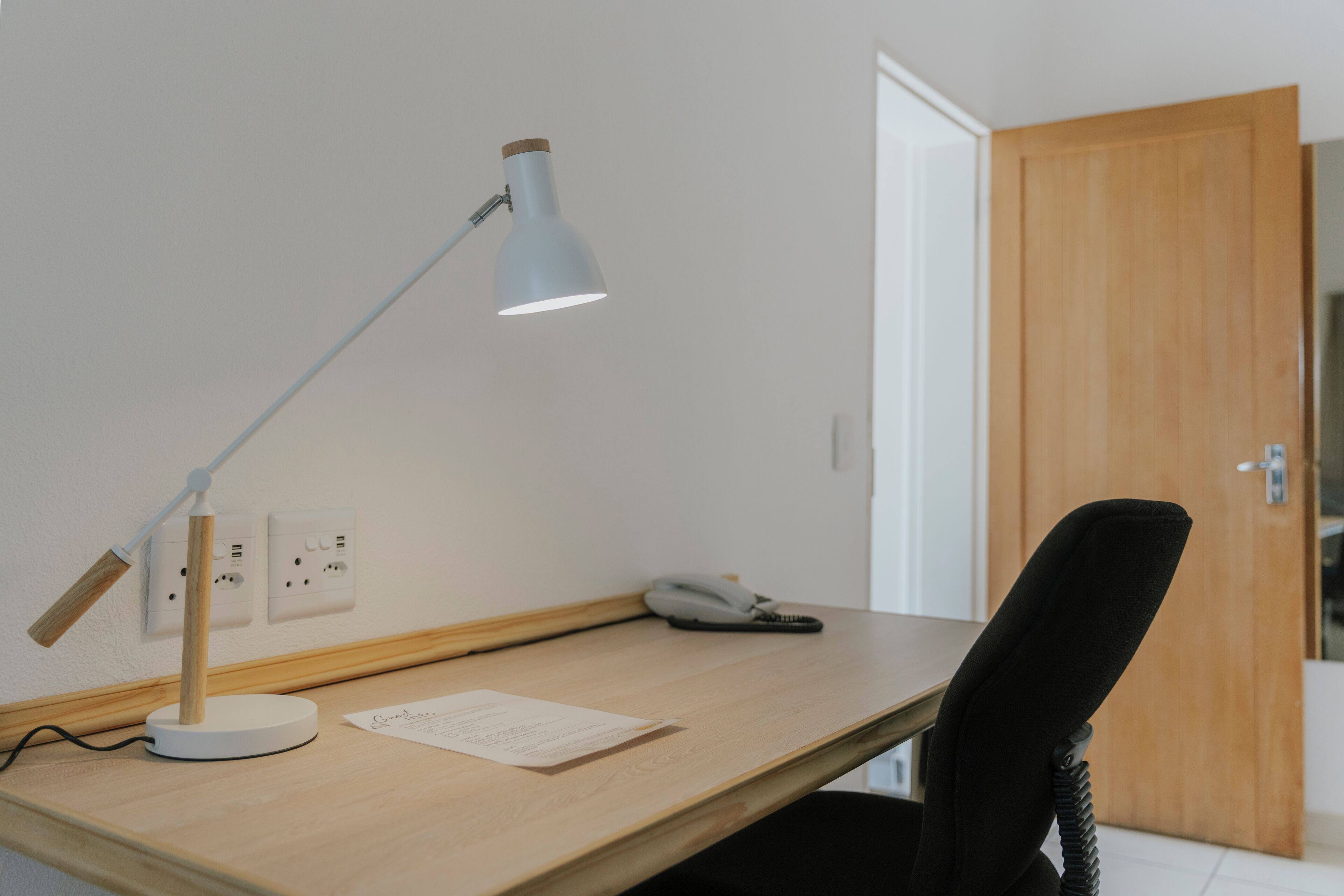 A wooden desk with a white desk lamp, a telephone, and a piece of paper.