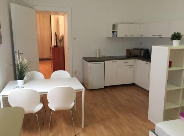 A compact kitchen with white cabinets, a countertop, and a small refrigerator. There is a dining area with a white table and chairs, and a potted plant on the table.