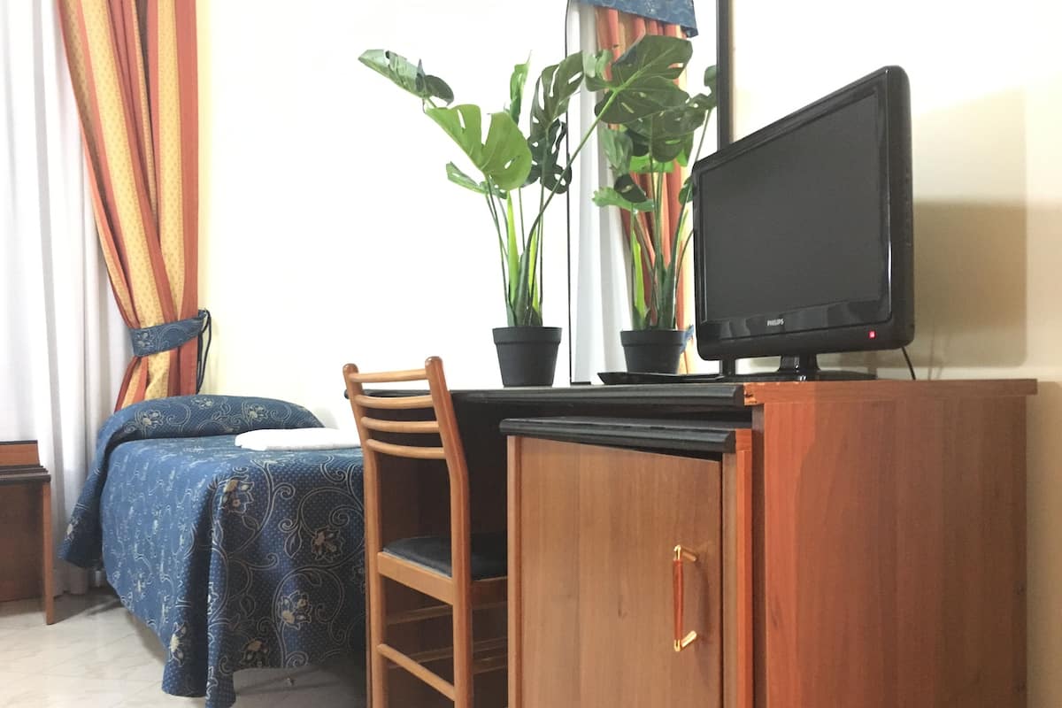 Quarto com uma cama, uma escrivaninha com computador, uma cadeira e duas plantas em vasos.