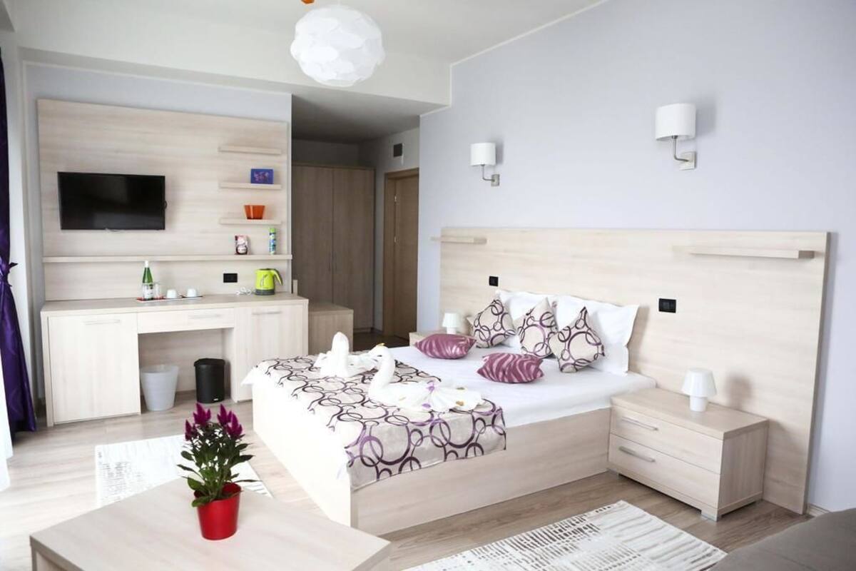 A modern bedroom with a wooden headboard, a flat-screen TV mounted above a desk, a bedside table with a lamp, and a decorative swan on the bed.