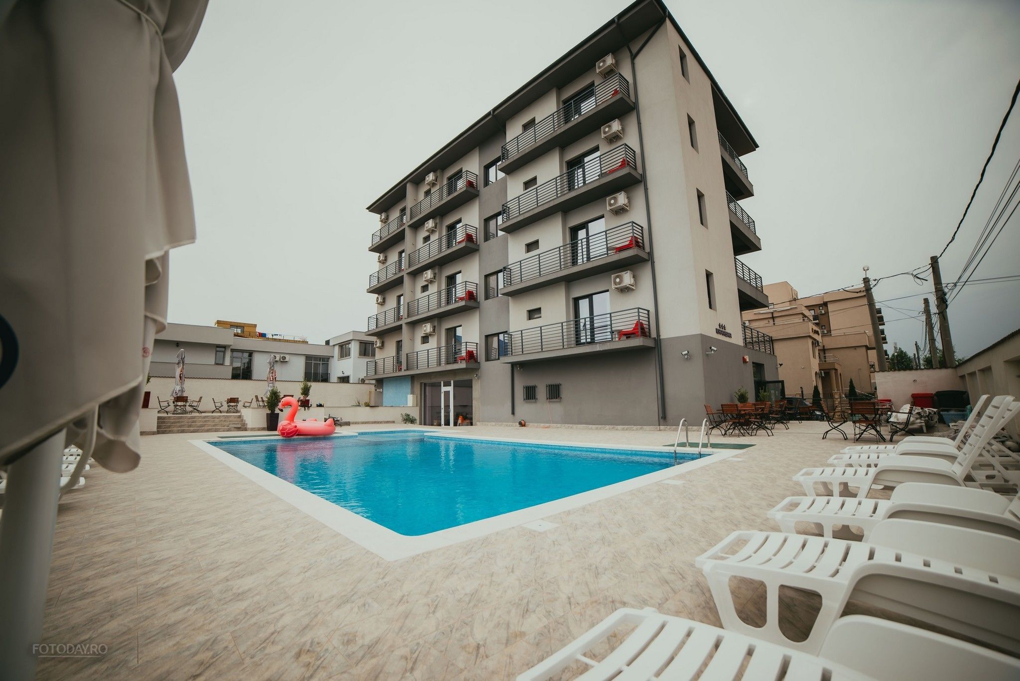 A swimming pool with lounge chairs, a pink flamingo float, and a multi-story building with balconies.