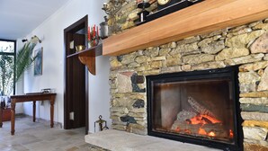 Una chimenea de piedra con repisa de madera, una mesa de comedor y una planta en maceta en una habitación con suelo de piedra.